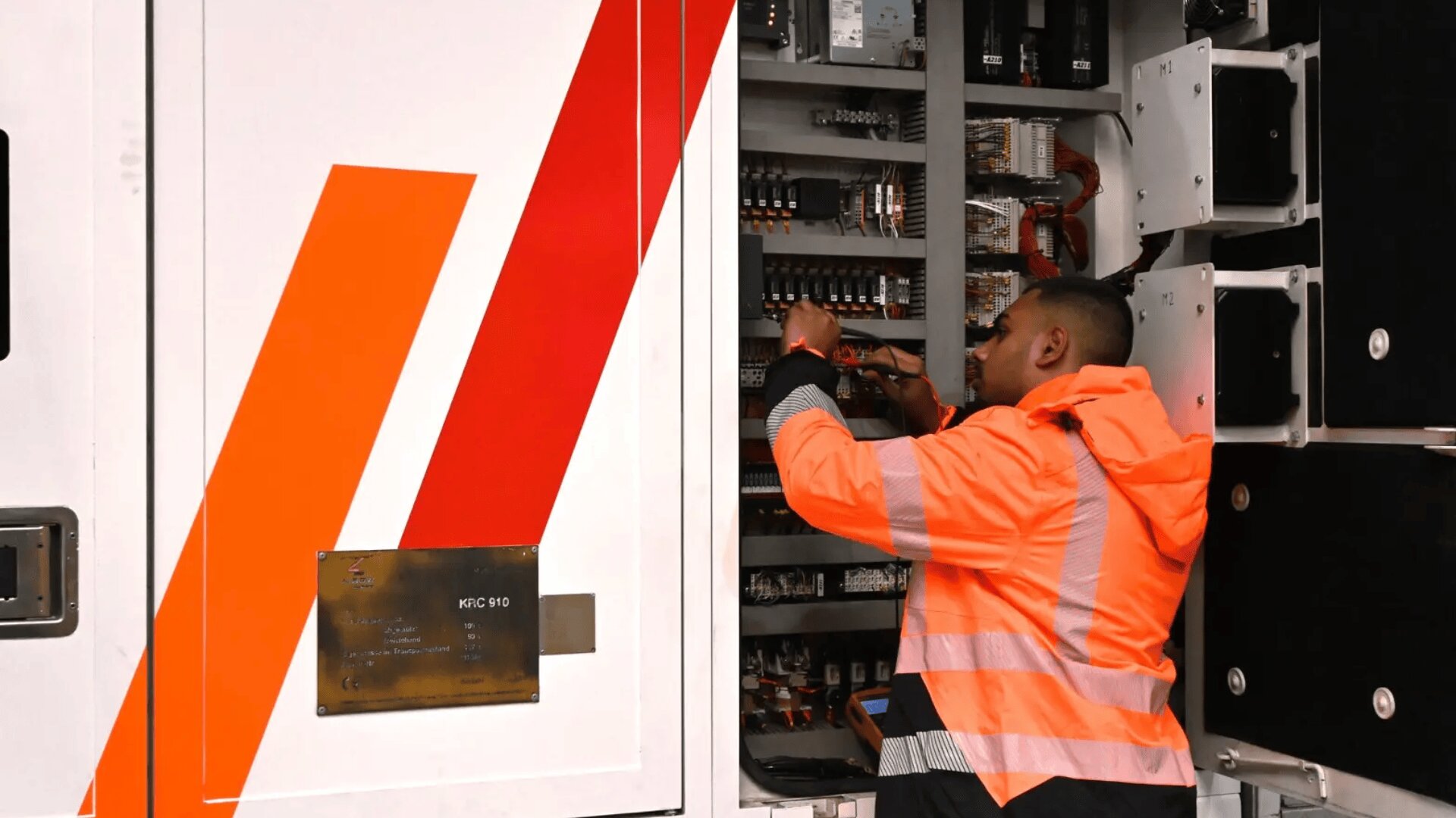 Eine Person in einer orangefarbenen Warnweste arbeitet mit einem Schraubenzieher an elektrischen Bauteilen in einem offenen industriellen Schaltschrank. Die Tür der Schalttafel ist mit roten und orangefarbenen Diagonalstreifen versehen.