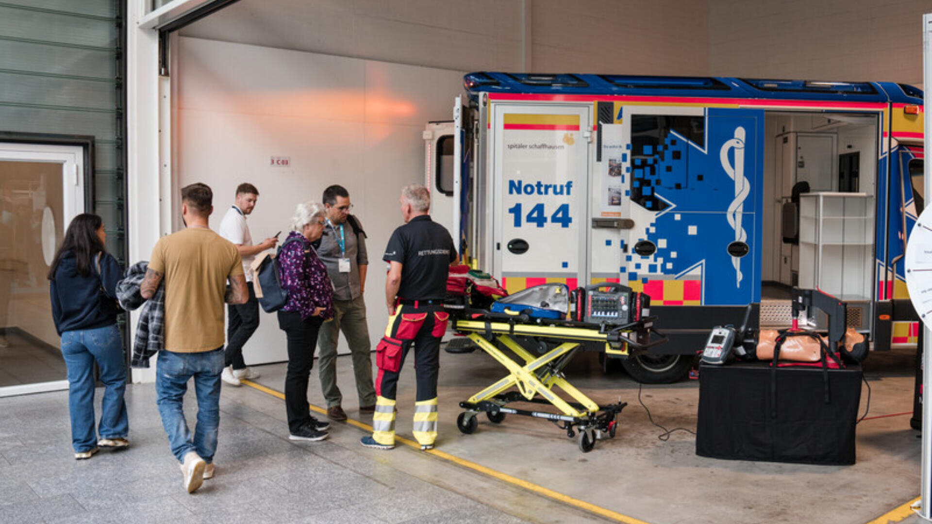 Eine Gruppe von Menschen steht in der Nähe eines Sanitäters und eines offenen Krankenwagens mit Notfallausrüstung, darunter eine Trage und eine HLW-Attrappe, in einem garagenähnlichen Raum.