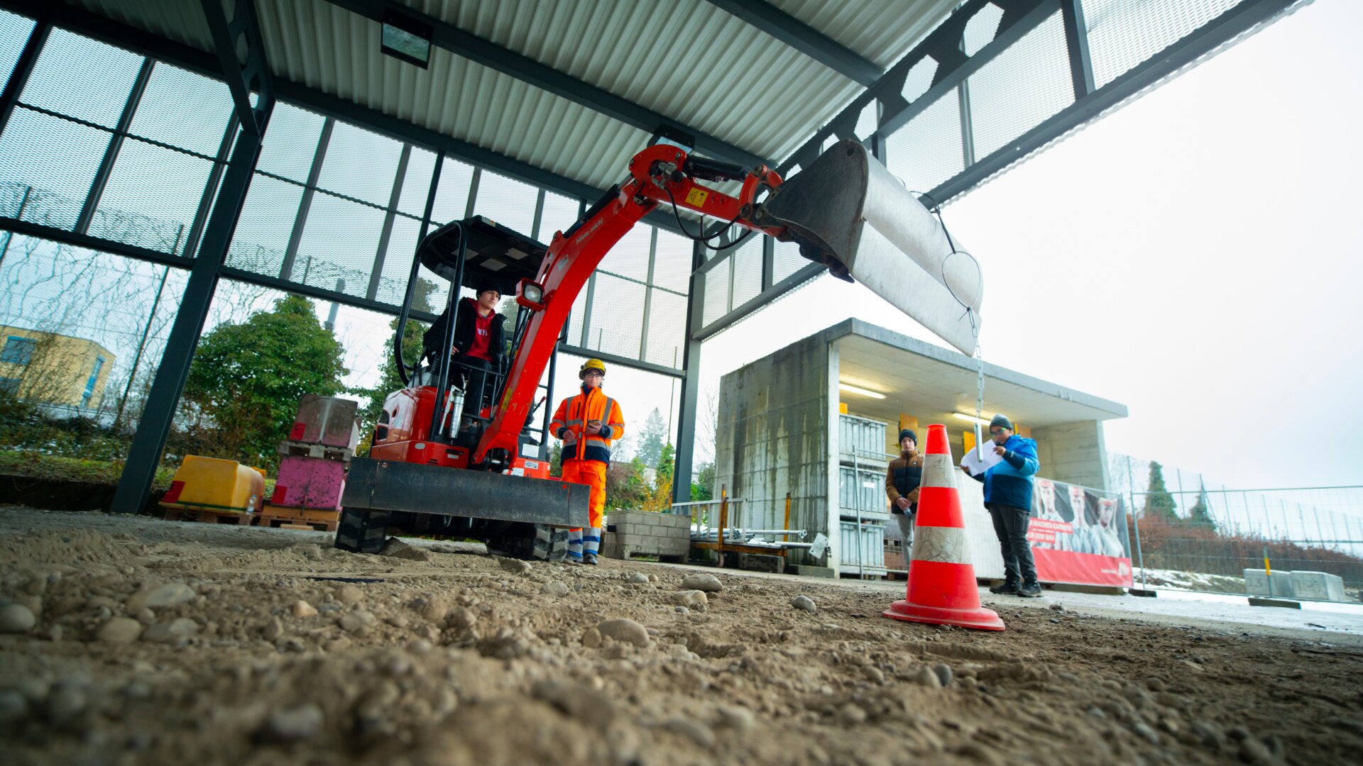 Ein kleiner roter Bagger, der von einem Arbeiter bedient wird, hebt eine Schaufel über Verkehrsleitkegel in einer teilweise geöffneten Baustelle, während zwei andere Arbeiter in Schutzausrüstung den Vorgang beobachten und anleiten.