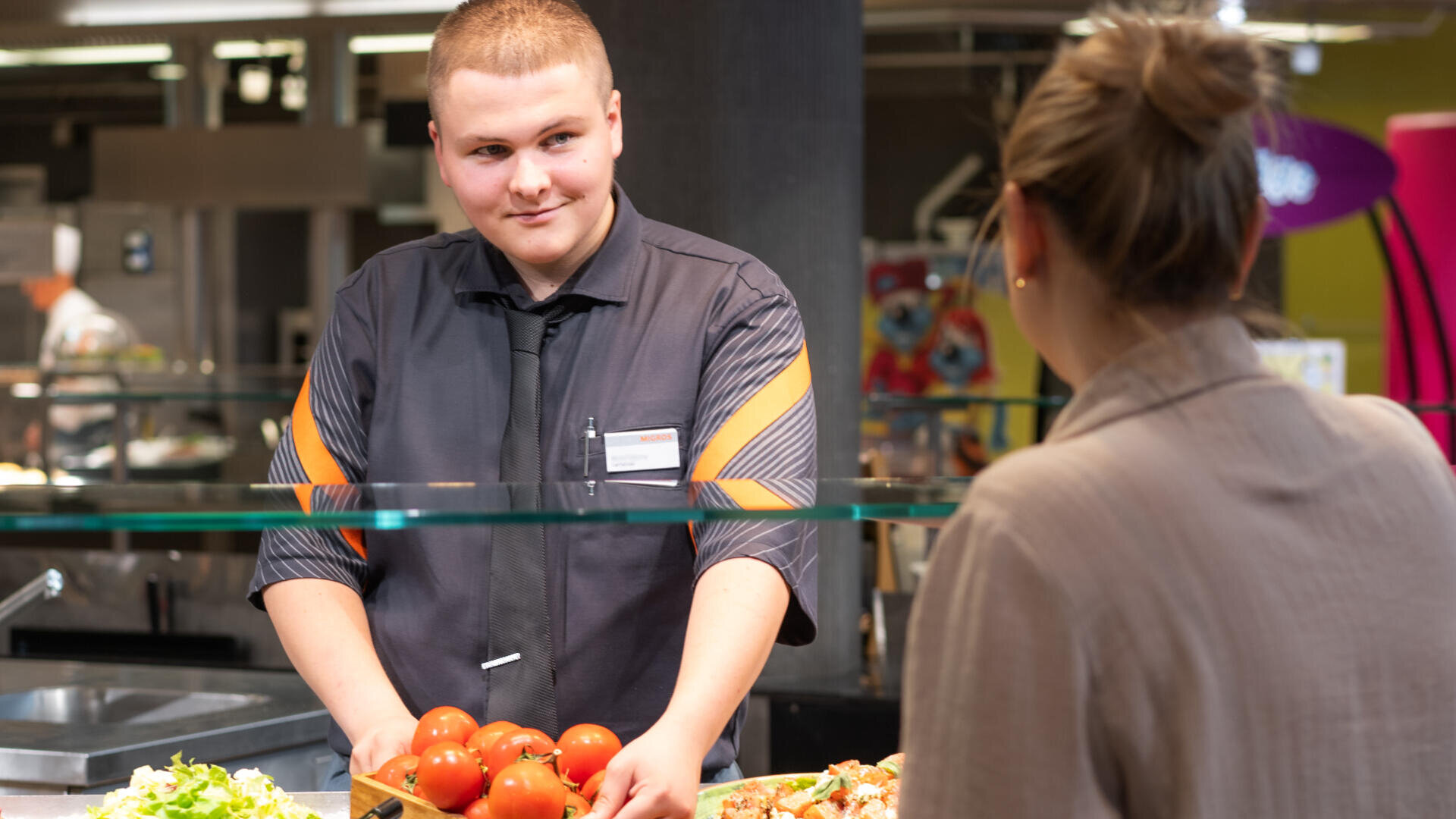 Ein Kantinenangestellter in Uniform steht hinter einer Theke mit Tomaten in der Hand, lächelt und spricht mit einer Kundin, die auf der anderen Seite steht. Auf der Theke liegen Salat und andere Lebensmittel.