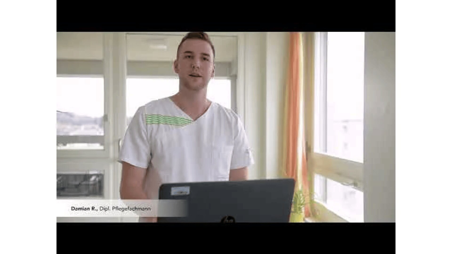 Ein Mann im Arztkittel steht hinter einem Laptop in einem hellen Raum mit großen Fenstern. In der Ecke steht ein Schild mit der Aufschrift Damian R., Dipl. Pflegefachmann.