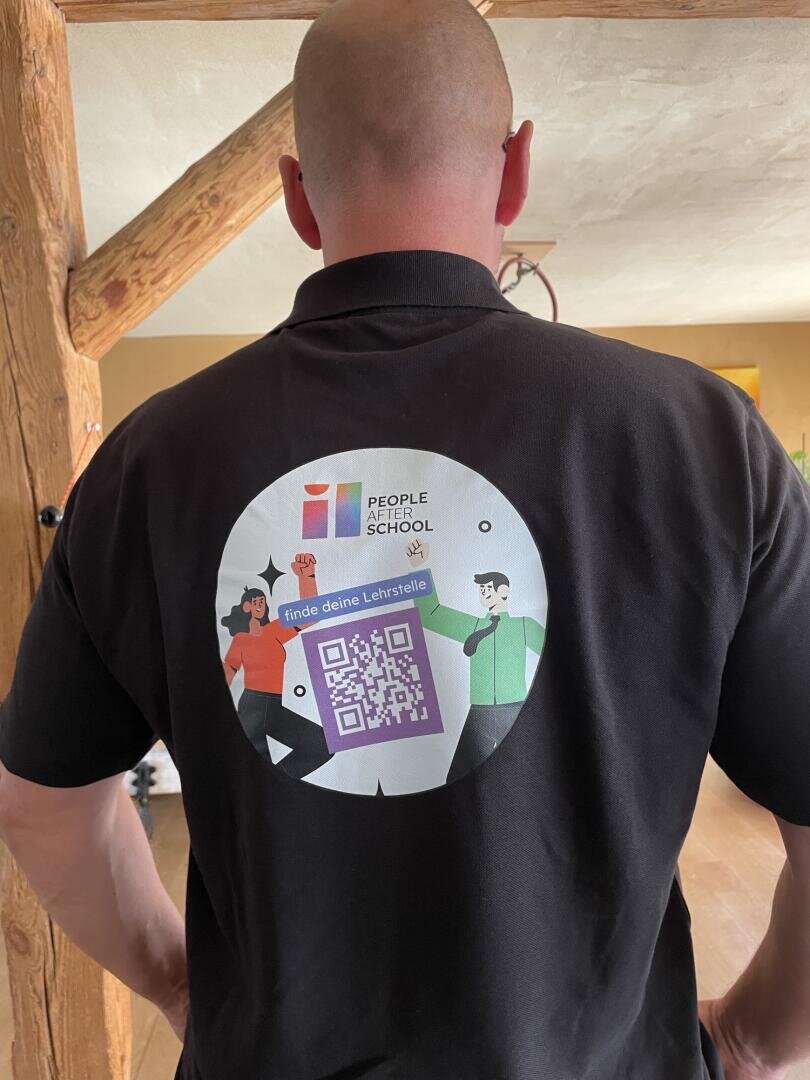 Eine Person, die ein schwarzes Hemd mit einem großen kreisförmigen Logo auf dem Rücken trägt, das Cartoon-Figuren, die Aufschrift People School und einen violetten QR-Code, eingerahmt von roten und grünen Akzenten, zeigt.