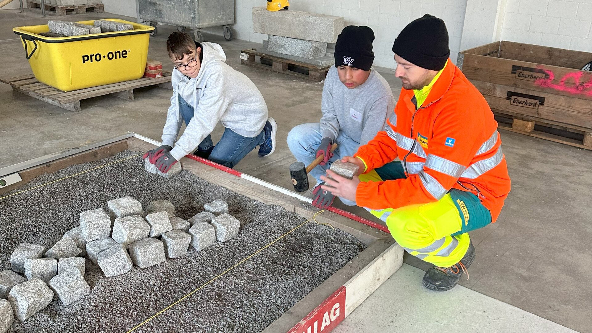 Drei Personen, zwei in Freizeitkleidung und eine in einer leuchtend orange-gelben Arbeitsuniform, knien neben einer mit Kies und Steinblöcken gefüllten Grube und ordnen Steine als Teil einer Bau- oder Trainingsaktivität in einem Gebäude an.