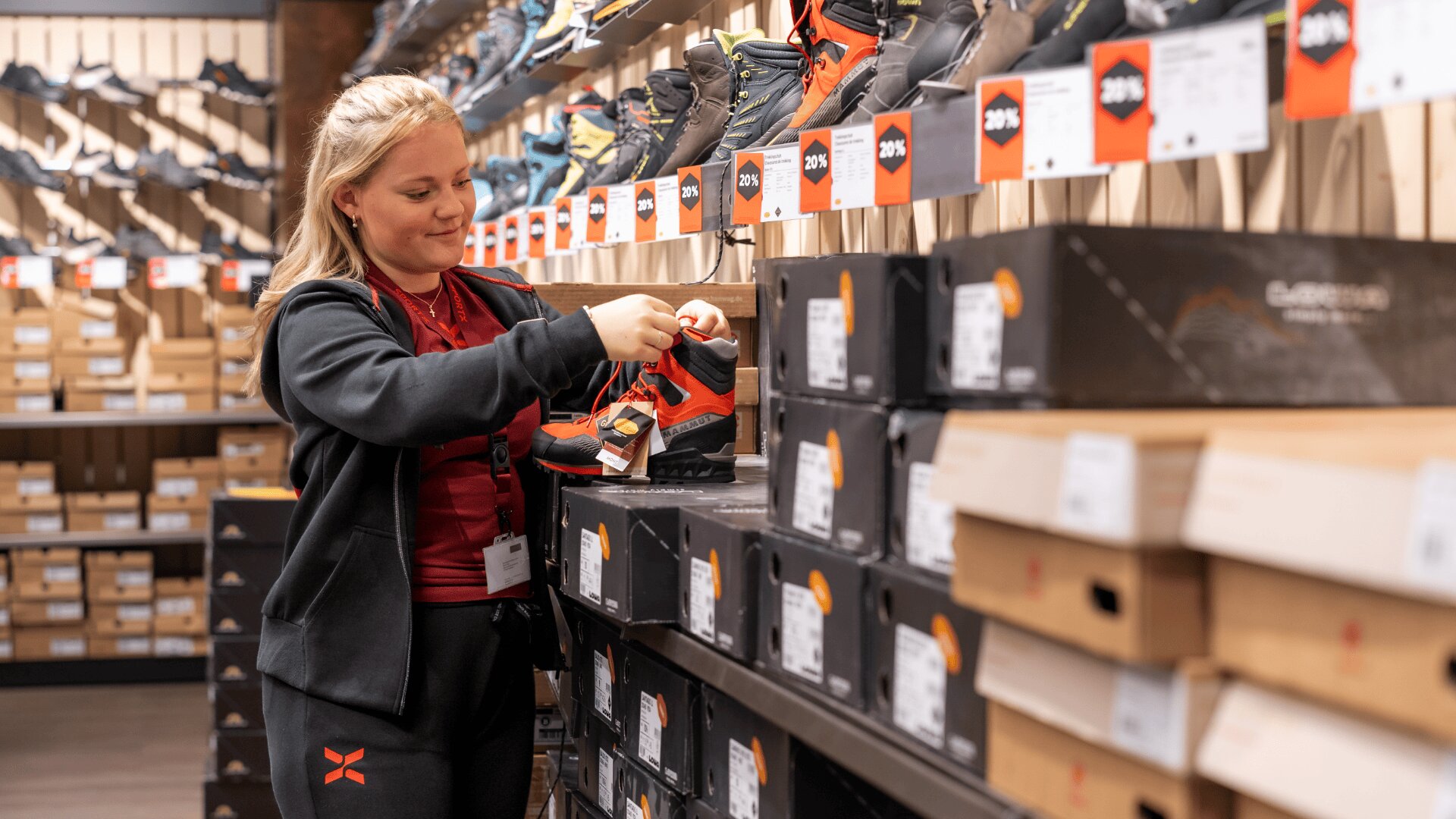 Eine Frau in einem Sportbekleidungsgeschäft begutachtet einen orange-schwarzen Schuh, umgeben von Regalen und Kartons mit Schuhen. Auf Schildern über den Schuhen sind Preisnachlässe angegeben.