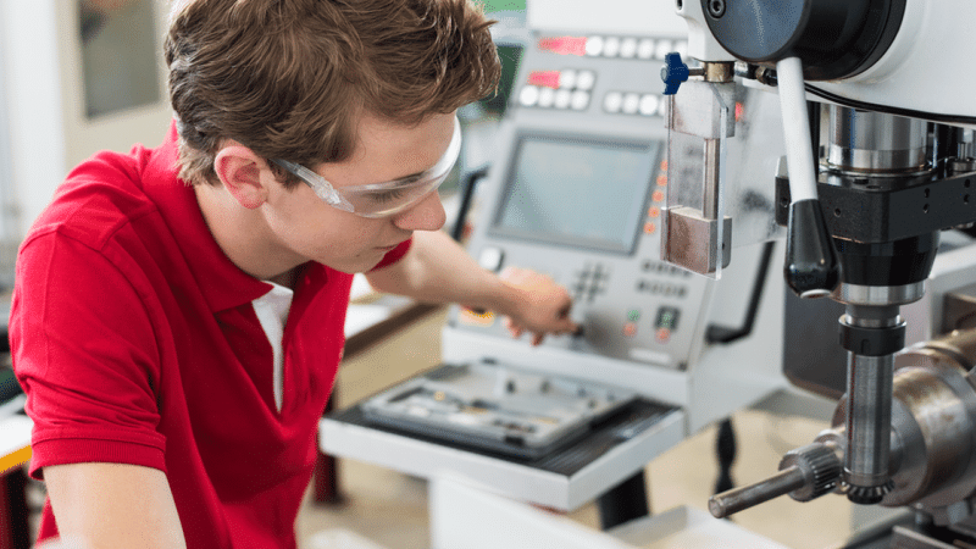 Ein junger Mensch, der eine Schutzbrille und ein rotes Hemd trägt, bedient Maschinen in einer Werkstatt oder einem Labor und konzentriert sich auf die Bedienelemente und den Monitor einer komplexen Industriemaschine.