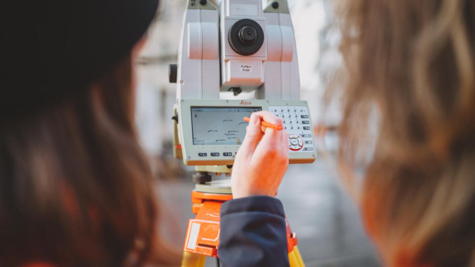 Zwei Personen stehen in der Nähe eines Vermessungsinstruments, während eine Person mit einem Stift das digitale Bedienfeld bedient und sich auf Messungen oder Dateneingaben im Freien konzentriert.