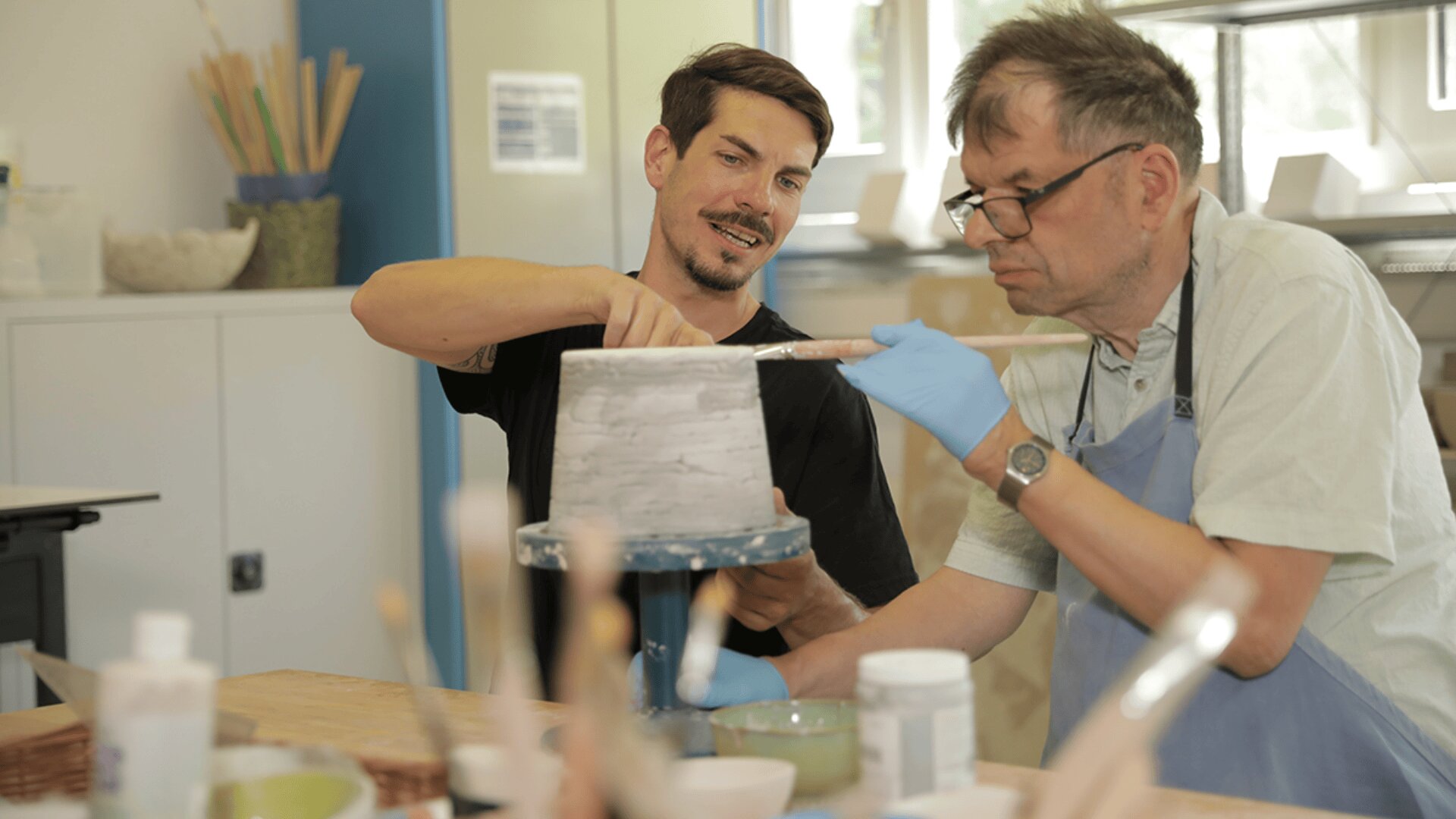 Zwei Männer sitzen an einem Tisch und bemalen gemeinsam ein Töpferstück. Ein Mann, der ein schwarzes Hemd trägt, lächelt und führt den anderen, der eine Brille, Handschuhe und eine Schürze trägt, beim Auftragen von Farbe mit Pinseln in einem hellen Kunstatelier.