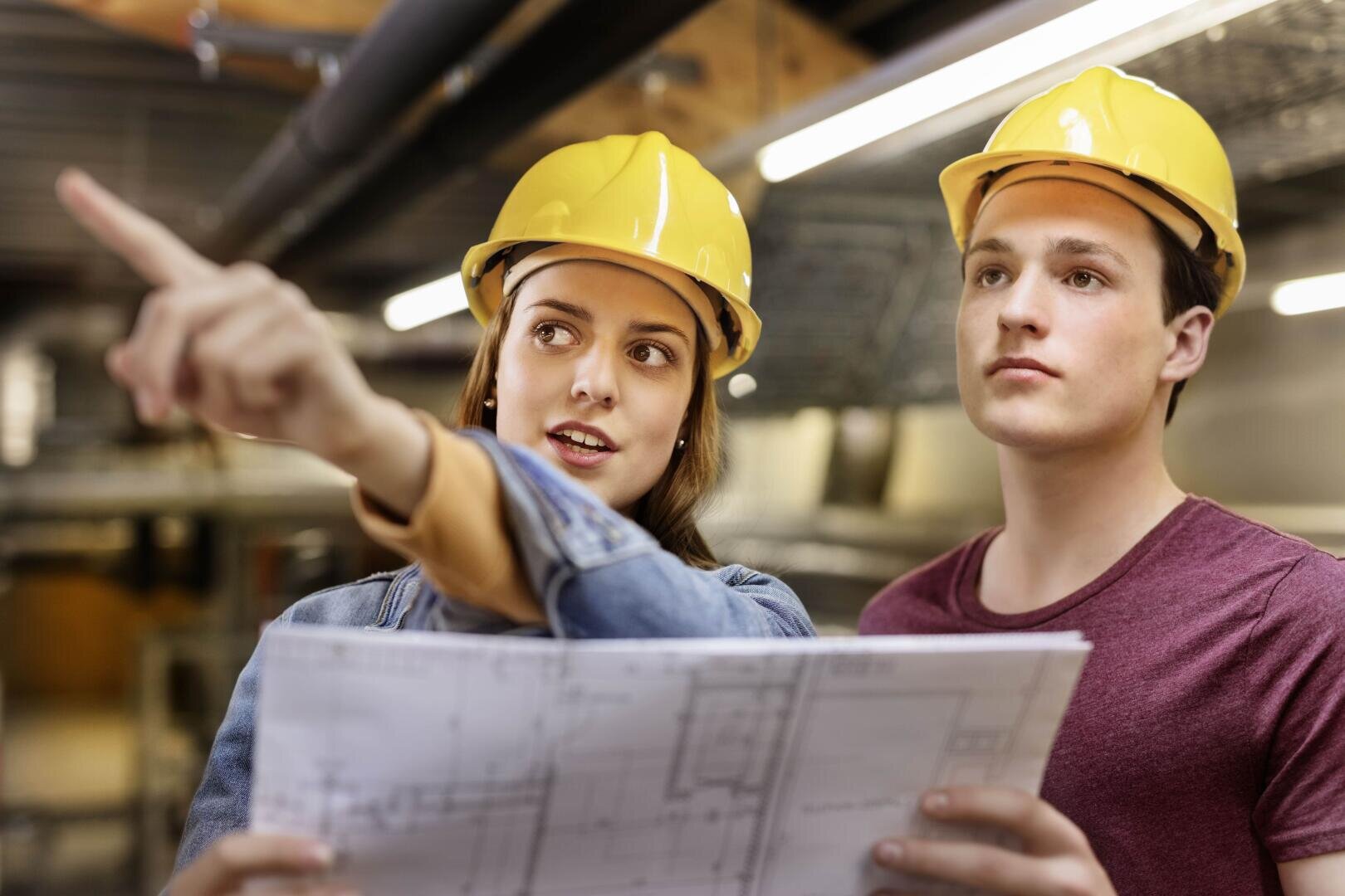 Zwei junge Erwachsene mit gelben Schutzhelmen stehen in einem Haus; einer hält einen Bauplan in der Hand, während der andere nach vorne zeigt und scheinbar über Bau- oder Renovierungspläne in einer Industrie- oder Bauumgebung spricht.
