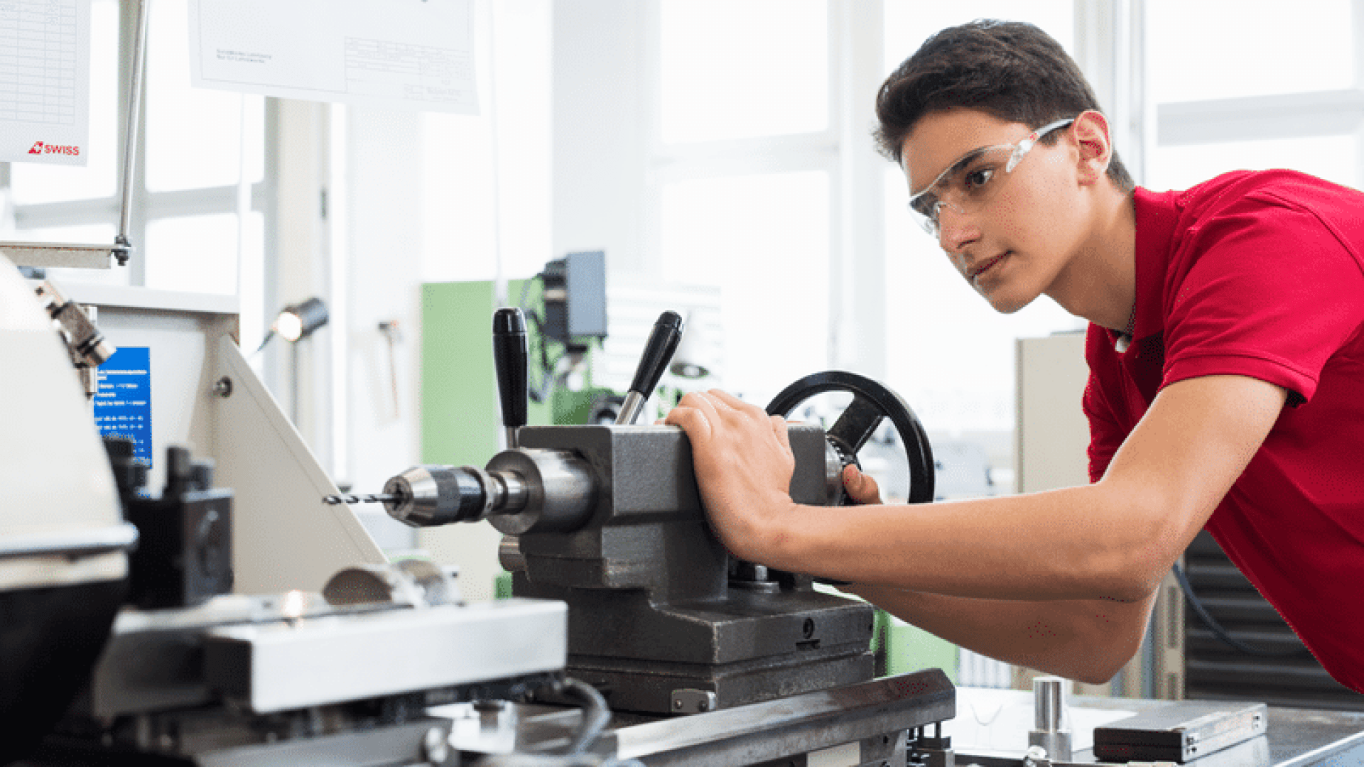 Ein junger Mann mit Schutzbrille und rotem Hemd bedient in einer hellen, modernen Werkstatt eine Drehmaschine und ist konzentriert mit der Einstellung der Geräte beschäftigt.