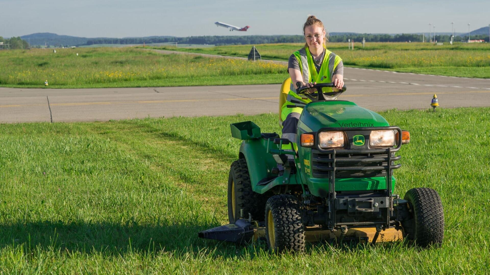 Eine Person, die eine gelbe Sicherheitsweste trägt, fährt mit einem grünen Aufsitzrasenmäher auf Gras in der Nähe einer Landebahn eines Flughafens, während im Hintergrund ein Flugzeug abhebt.