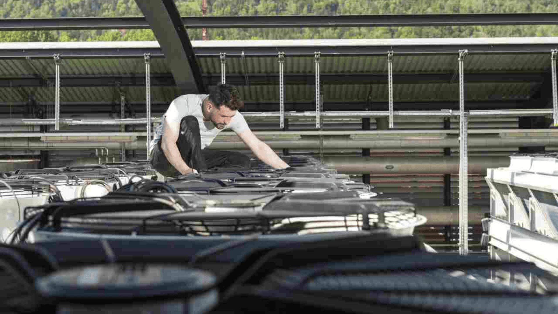 Ein Mann in einem weißen T-Shirt hockt auf einer großen Industrieanlage und inspiziert oder arbeitet an den Geräten im Freien, im Hintergrund sind Bäume und Metallstrukturen zu sehen.