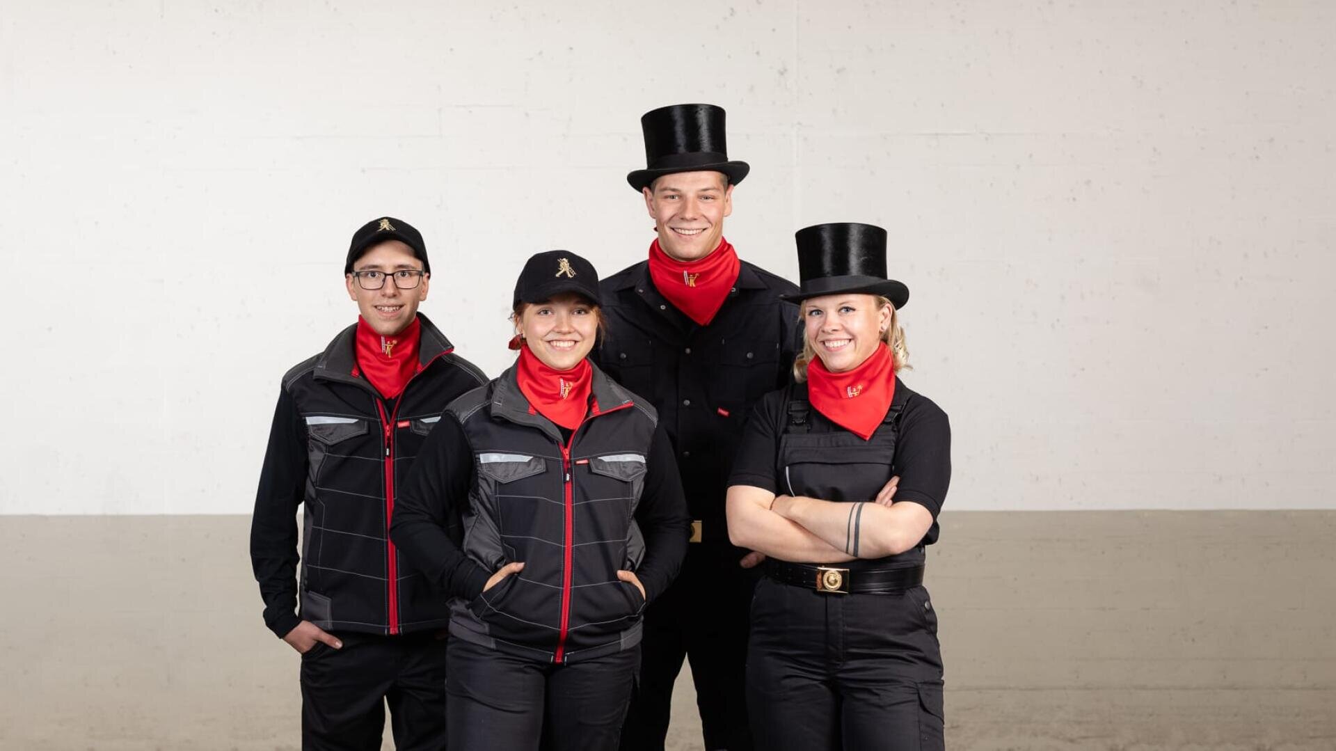 Vier Personen in schwarzen Uniformen, roten Halstüchern und Hüten - zwei mit schwarzen Zylinderhüten und zwei mit schwarzen Baseballkappen - stehen vor einem schlichten, hellen Hintergrund zusammen.