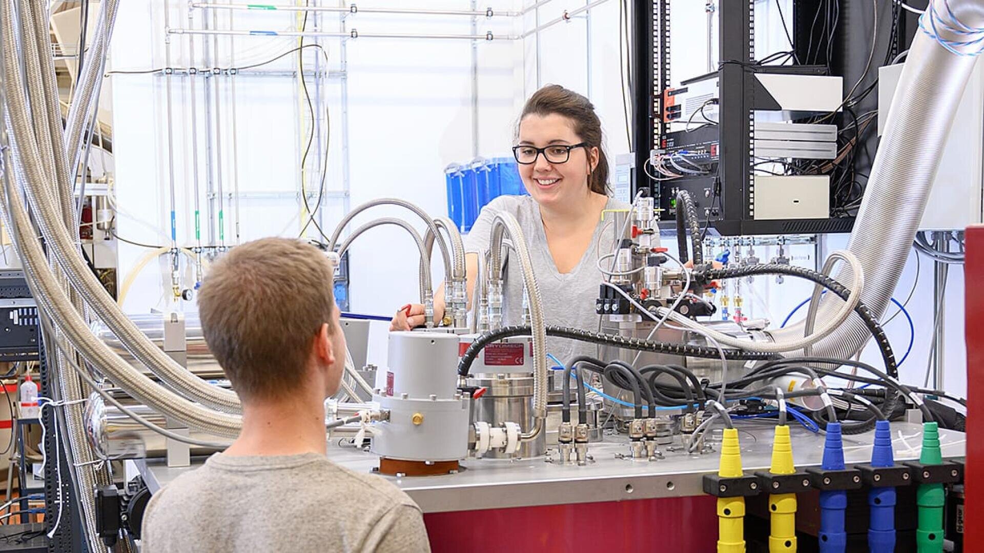 Eine Frau lächelt, während sie in einem Labor mit komplexen wissenschaftlichen Geräten und Röhren arbeitet und mit einem Mann spricht, der ihr gegenüber sitzt. Im Vordergrund sind bunte Laborgeräte und Drähte zu sehen.