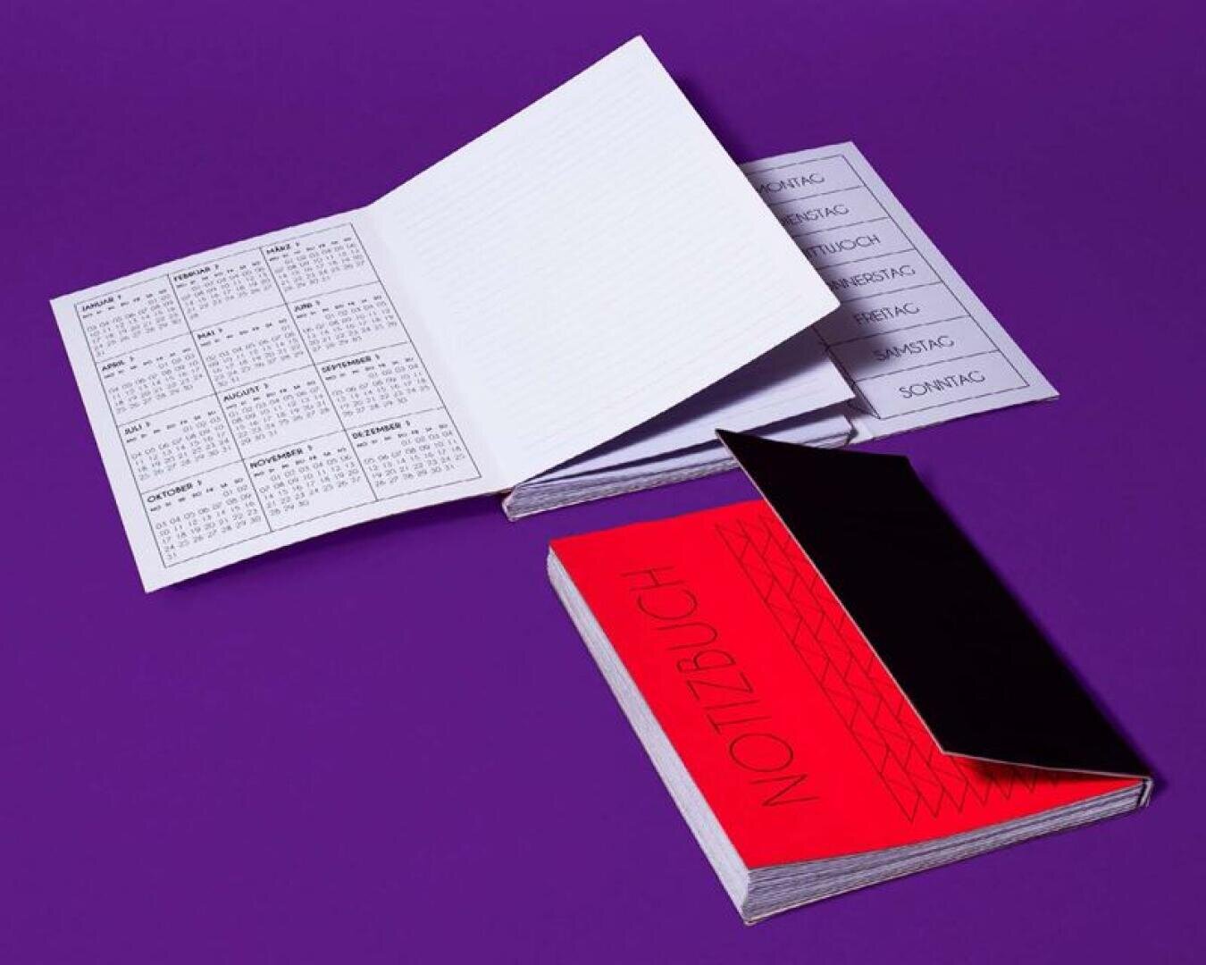 Zwei aufgeschlagene Notizbücher auf lilafarbener Oberfläche; eines zeigt einen Kalender und linierte Seiten, das andere offenbart einen leuchtend roten Einband mit der Aufschrift NOTIZBUCH in fetten Buchstaben, teilweise verdeckt durch eine schwarze Klappe.