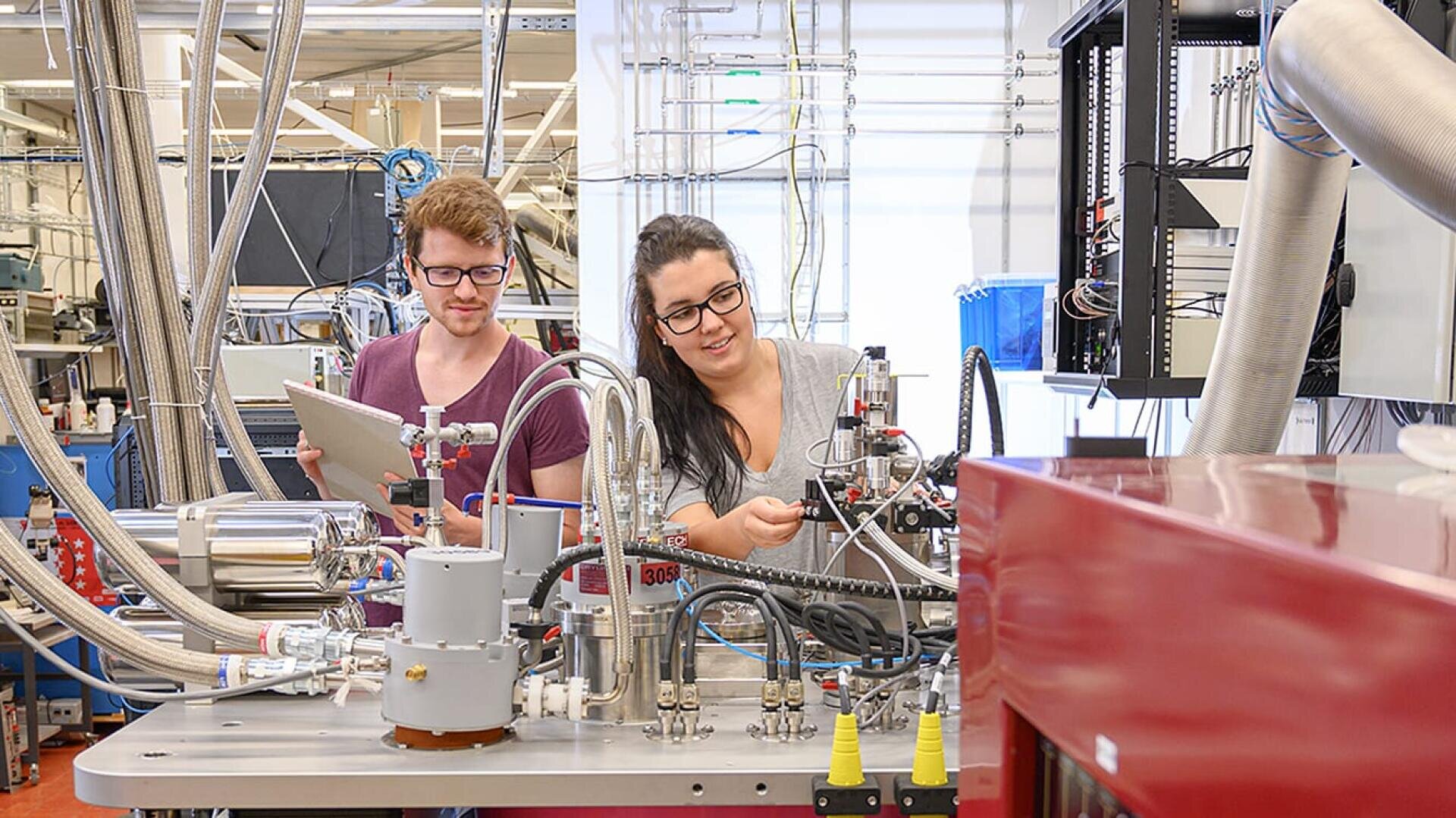 Zwei Brillenträger arbeiten gemeinsam in einem Labor und prüfen und justieren wissenschaftliche Geräte, die von Rohren, Kabeln und Maschinen umgeben sind. Eine Person hält ein Tablet, während die andere eine Maschine bedient.