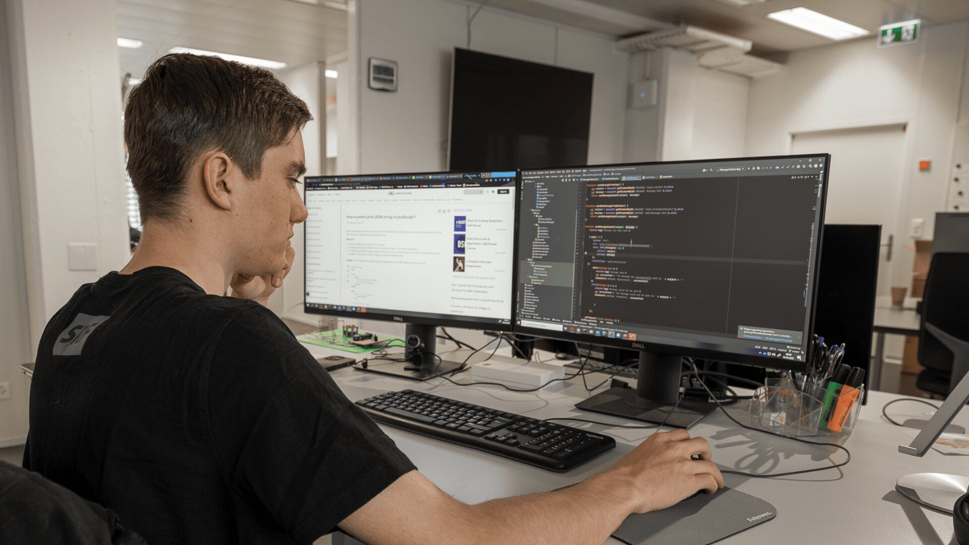 Eine Person sitzt an einem Schreibtisch und arbeitet an zwei Computermonitoren; ein Bildschirm zeigt einen Code, der andere eine Website an. Auf dem Schreibtisch in einer modernen Büroumgebung liegen Büromaterial und eine Tastatur.