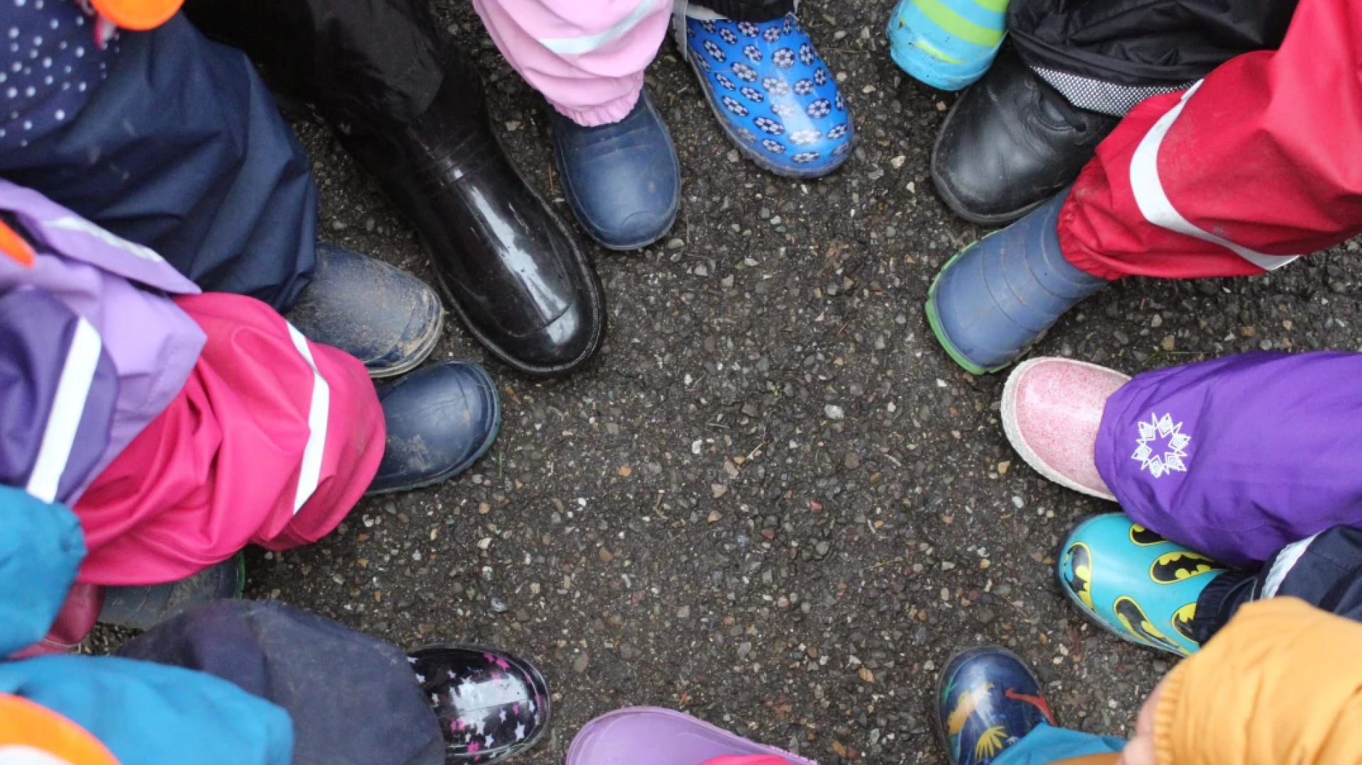 Eine Gruppe von Kindern, die in einem Kreis auf dem Asphalt stehen und nur ihre bunten Schuhe und Stiefel zeigen, bilden einen Ring um die leere Mitte.