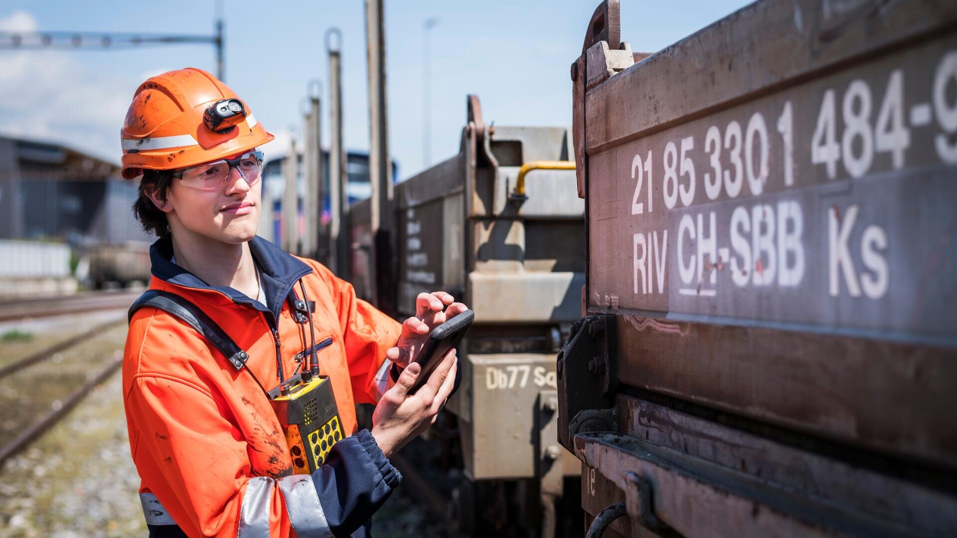 Ein Eisenbahner in orangefarbener Sicherheitsuniform und mit Helm benutzt ein Handheld-Gerät, um Informationen über einen Güterzugwaggon auf einem Rangierbahnhof zu prüfen und aufzuzeichnen.