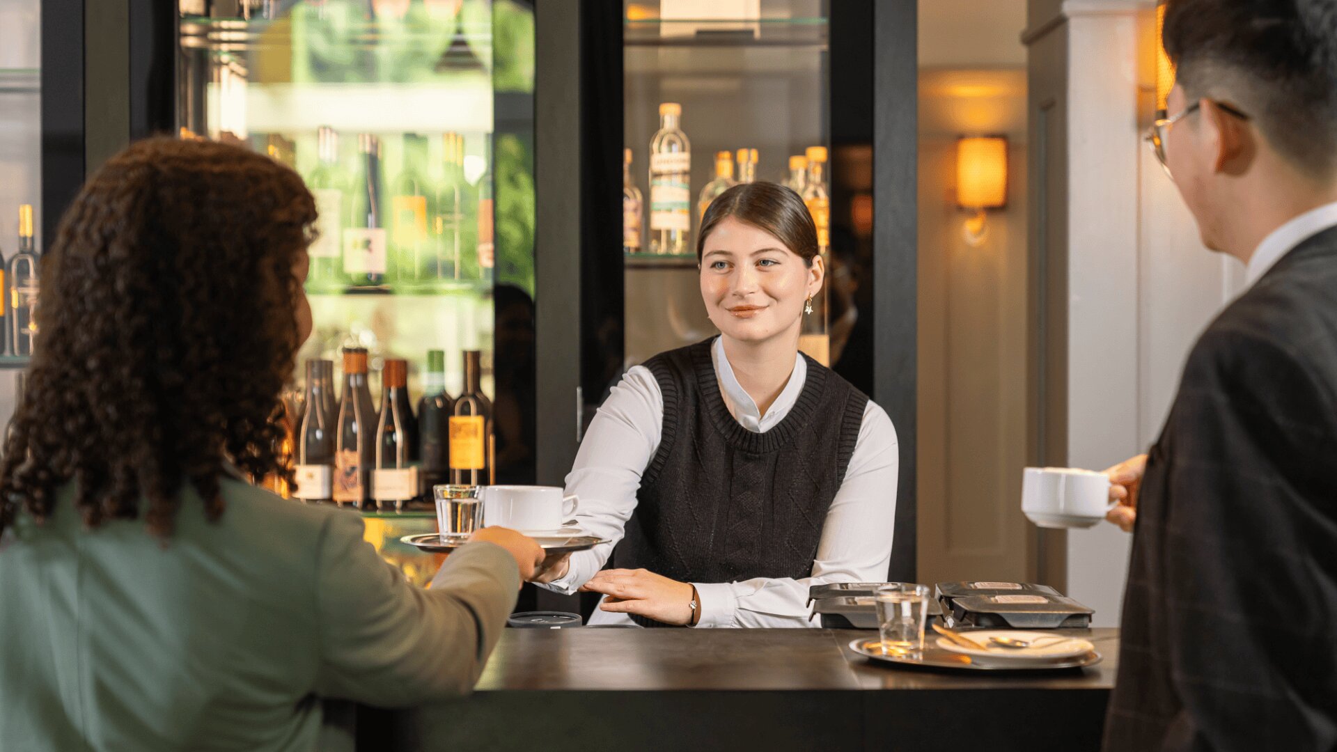 Eine Frau hinter der Theke serviert zwei Personen, die an der Theke sitzen, Kaffee und Wasser, während im Hintergrund Flaschen mit Spirituosen stehen. Die Atmosphäre wirkt professionell und freundlich.