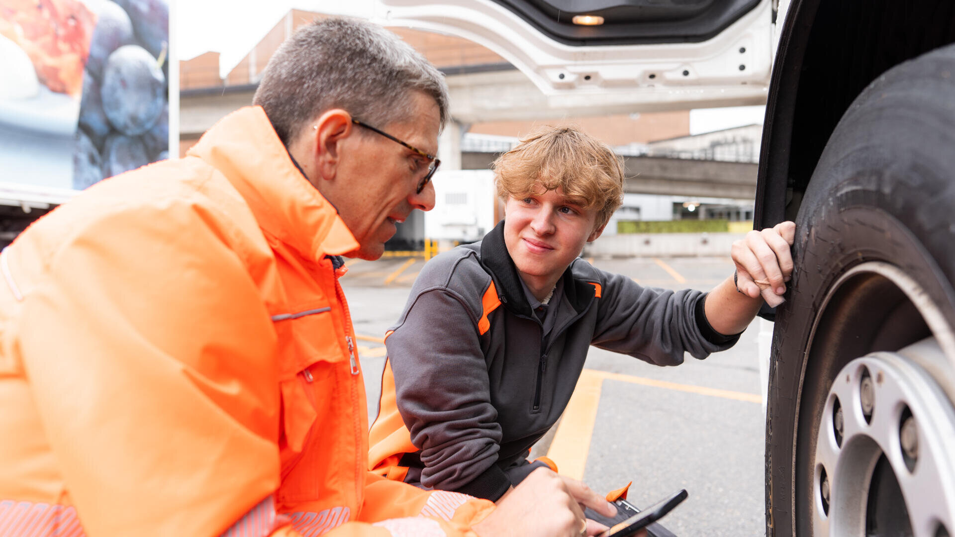 Zwei Männer in Arbeitsuniformen inspizieren einen großen Lkw-Reifen auf einem Parkplatz. Ein Mann hält ein Klemmbrett und einen Stift in der Hand, während der andere den Reifen berührt, während beide sich unterhalten.