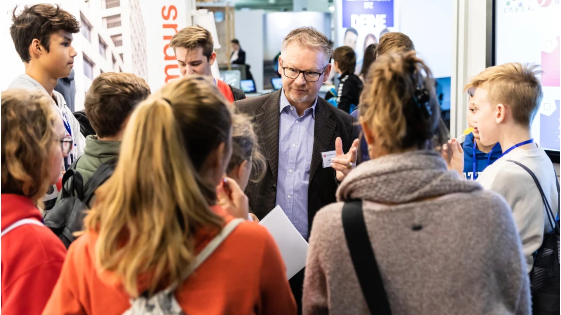 Ein Mann im Anzug spricht zu einer Gruppe Jugendlicher, die sich bei einer Veranstaltung oder Ausstellung in einer Halle um ihn herum versammelt haben, wobei im Hintergrund Messestände und Bildschirme zu sehen sind.