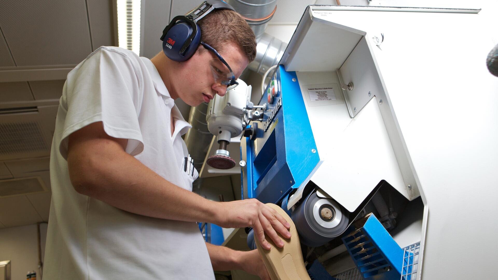 Eine Person mit Schutzbrille und Gehörschutz bedient eine Poliermaschine und bearbeitet einen beigen Gegenstand in einer Industrieumgebung.