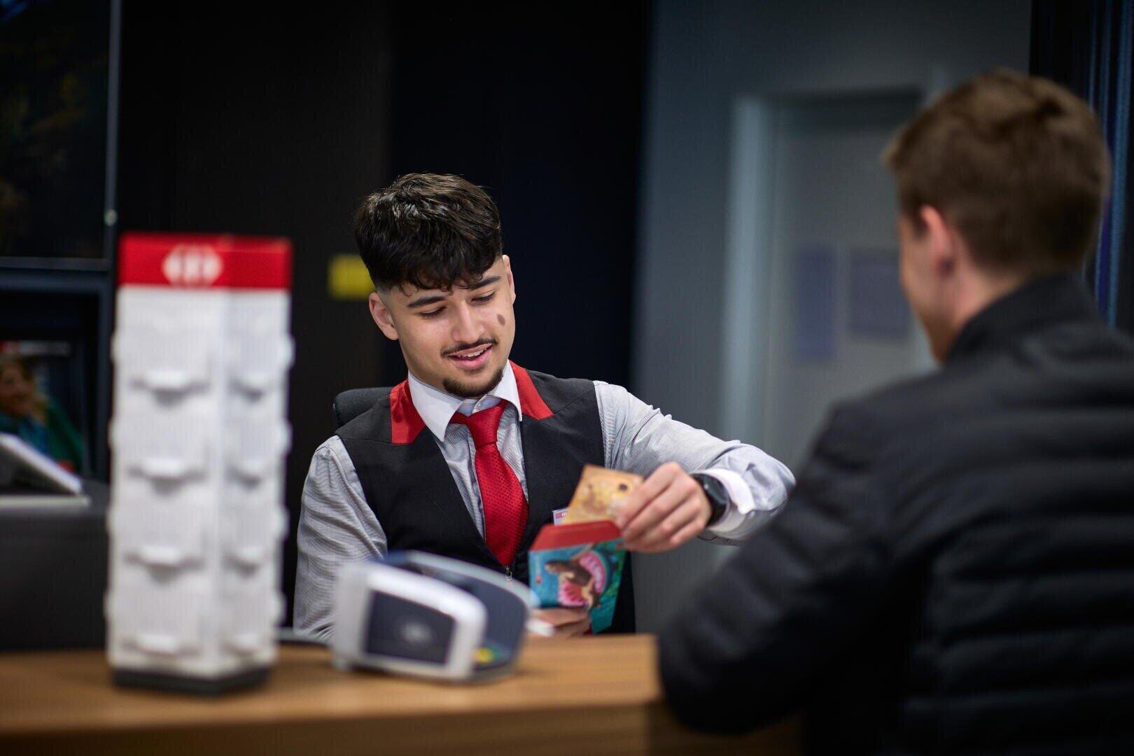 Ein Bankangestellter mit Weste und roter Krawatte hilft einem Kunden bei der Prüfung von Dokumenten oder Karten über einen Schalter in einer modernen Bank. Der Kunde ist teilweise sichtbar, von hinten gesehen.