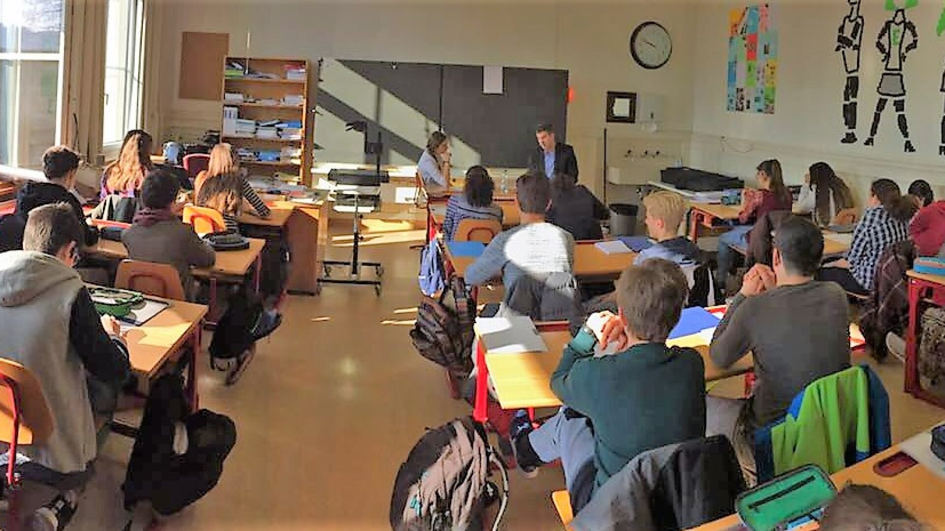 Ein Klassenzimmer mit Schülern, die an Tischen sitzen und einem Lehrer gegenübersitzen. Das Sonnenlicht fällt durch große Fenster ein, und im Raum sind Unterrichtsmaterialien und Regale zu sehen.