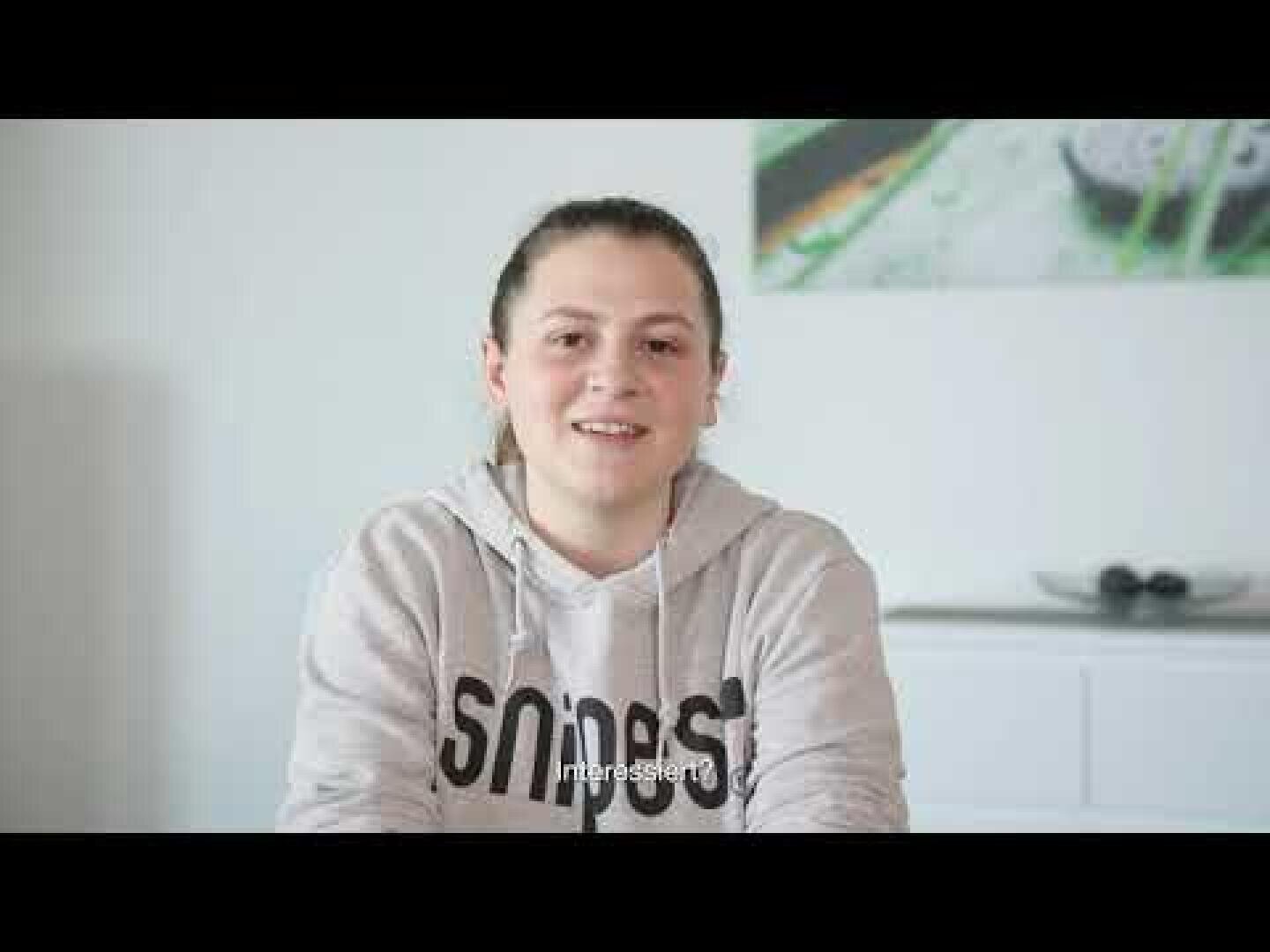 Eine Person mit heller Haut und zurückgebundenen braunen Haaren sitzt lächelnd in einem Haus und trägt einen hellen Kapuzenpulli mit Text darauf. Im Hintergrund befinden sich ein weißer Schrank und eine Wandmalerei.