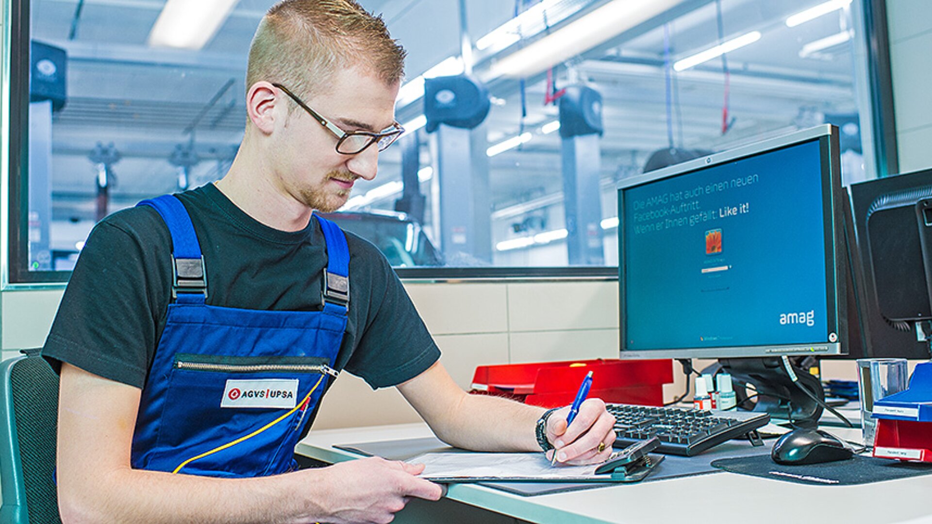 Ein Mann mit Brille und blauer Latzhose sitzt an einem Schreibtisch in einem Büro und schreibt auf einem Klemmbrett. Auf dem Schreibtisch befinden sich ein Computer, eine Tastatur und ein roter Schreibtisch-Organizer. Der Hintergrund zeigt einen hellen, modernen Arbeitsbereich.
