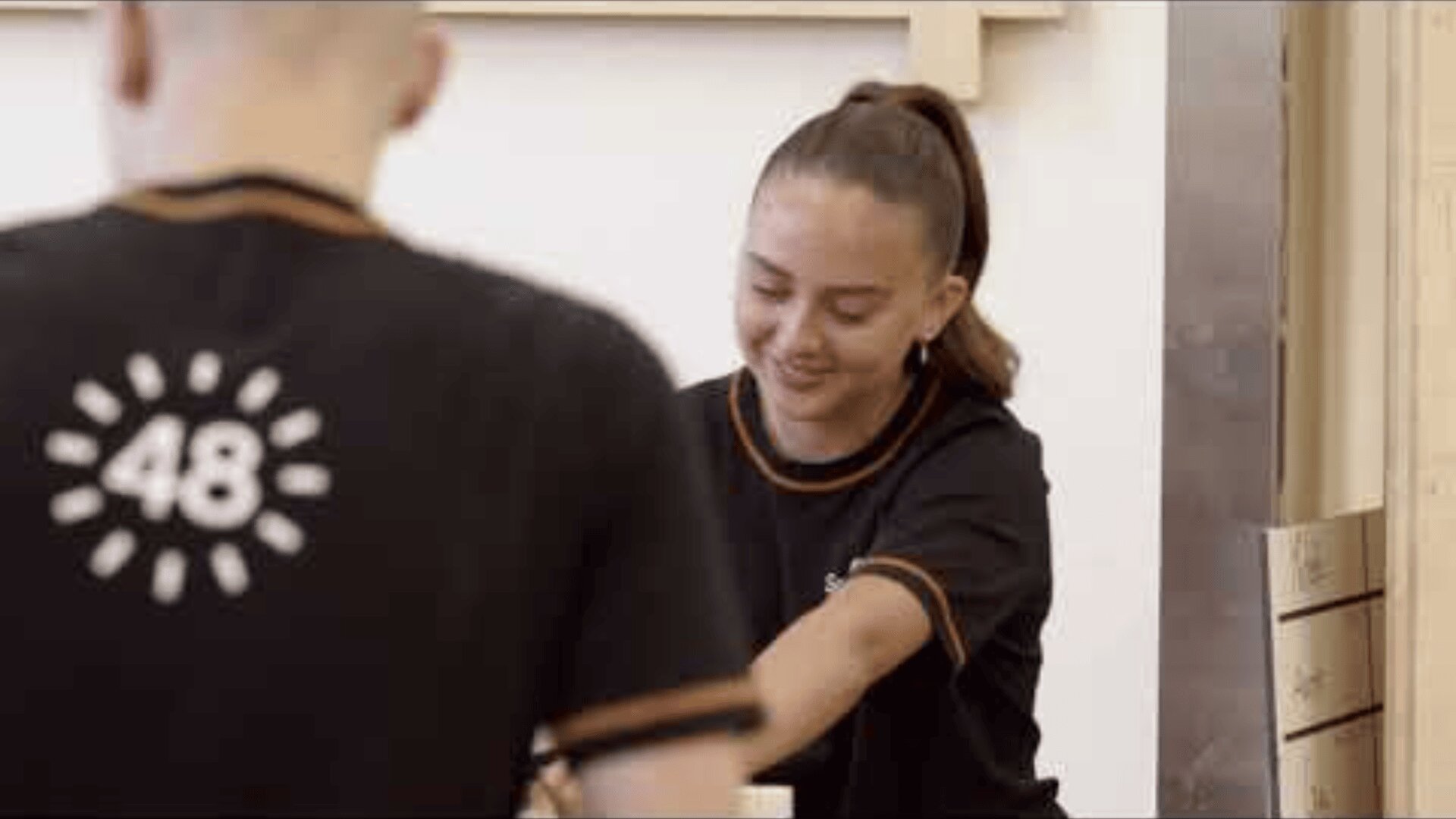 Eine junge Frau mit Pferdeschwanz lächelt, während sie neben einer Person arbeitet, die ein schwarzes T-Shirt mit der Nummer 48 und einer Sonnengrafik auf dem Rücken trägt. Sie scheinen in einem Innenraum an einer gemeinschaftlichen Aufgabe zu arbeiten.