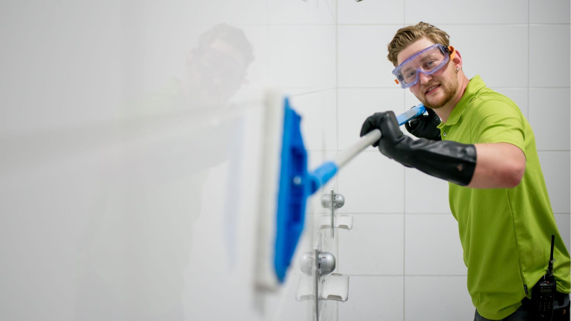 Eine Person, die eine Schutzbrille, schwarze Handschuhe und ein grünes Hemd trägt, reinigt lächelnd eine weiß geflieste Wand mit einem langstieligen blauen Mopp in einem Badezimmer.