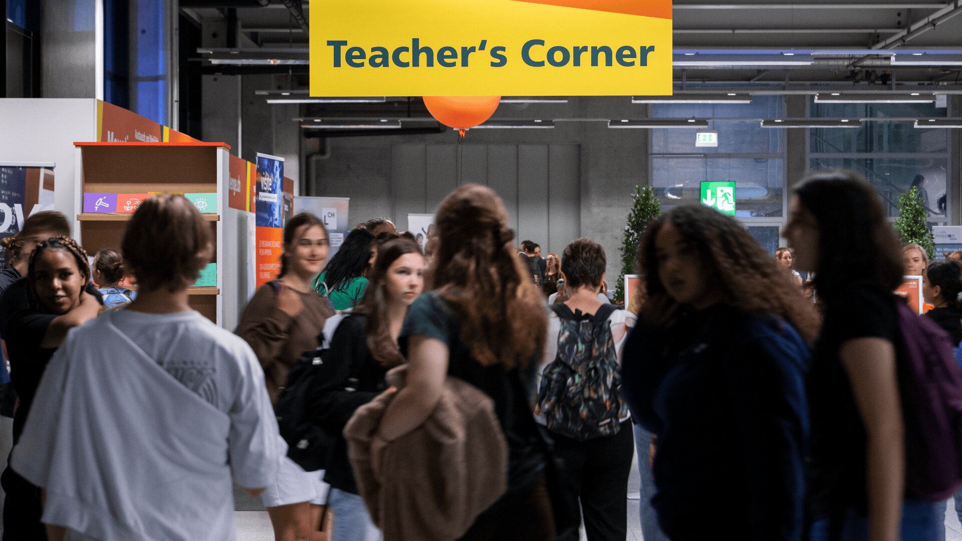 Eine Gruppe von Menschen geht und versammelt sich unter einem leuchtend gelben Teachers Corner-Schild in einem modernen, gut beleuchteten Innenraum mit Bücherregalen und Pflanzen im Hintergrund.