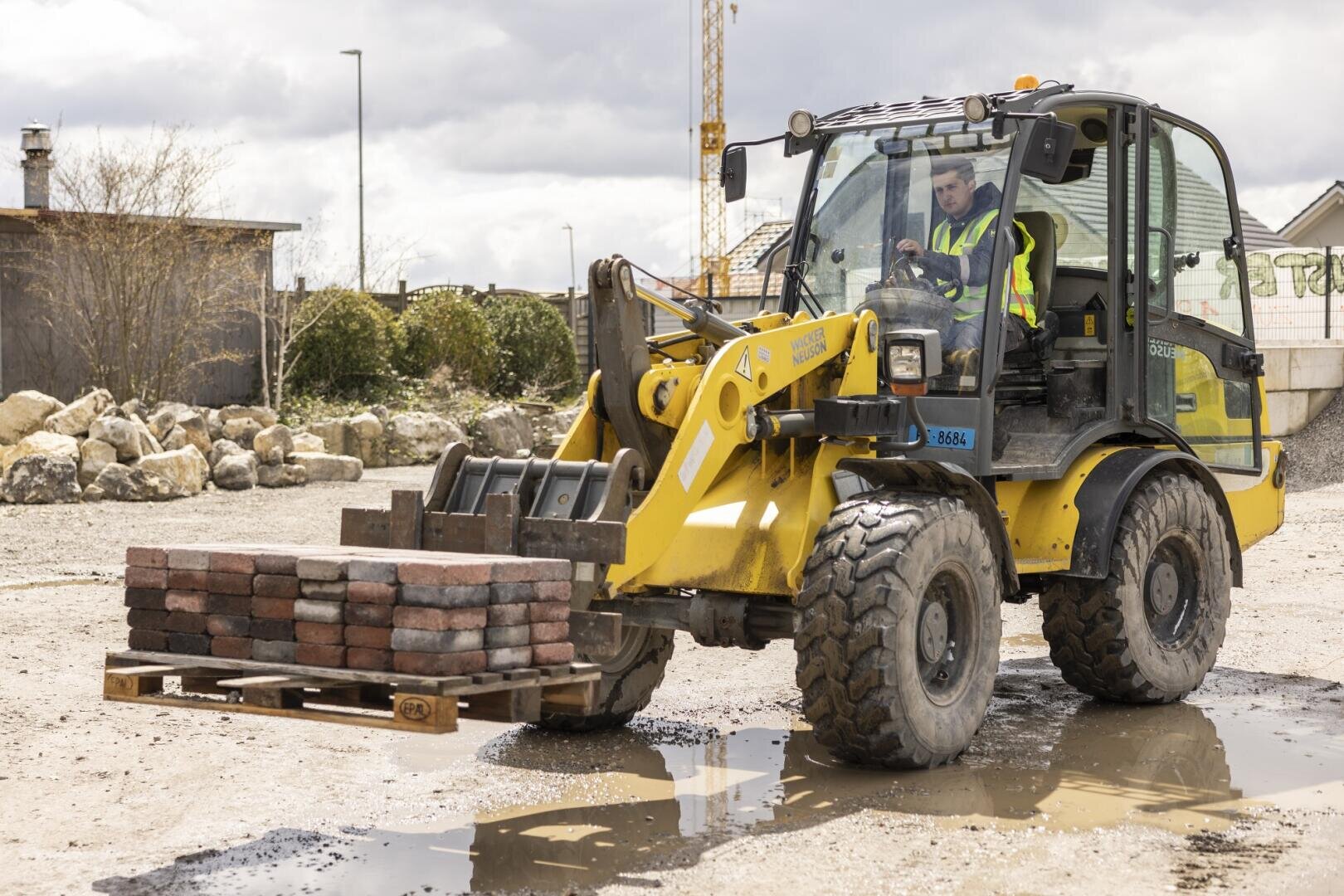Ein Bauarbeiter bedient einen gelben Frontlader und hebt eine Palette mit Ziegelsteinen auf einer Baustelle an. Der Boden ist nass und voller Pfützen, und im Hintergrund sind Felsen und Gebäude zu sehen.