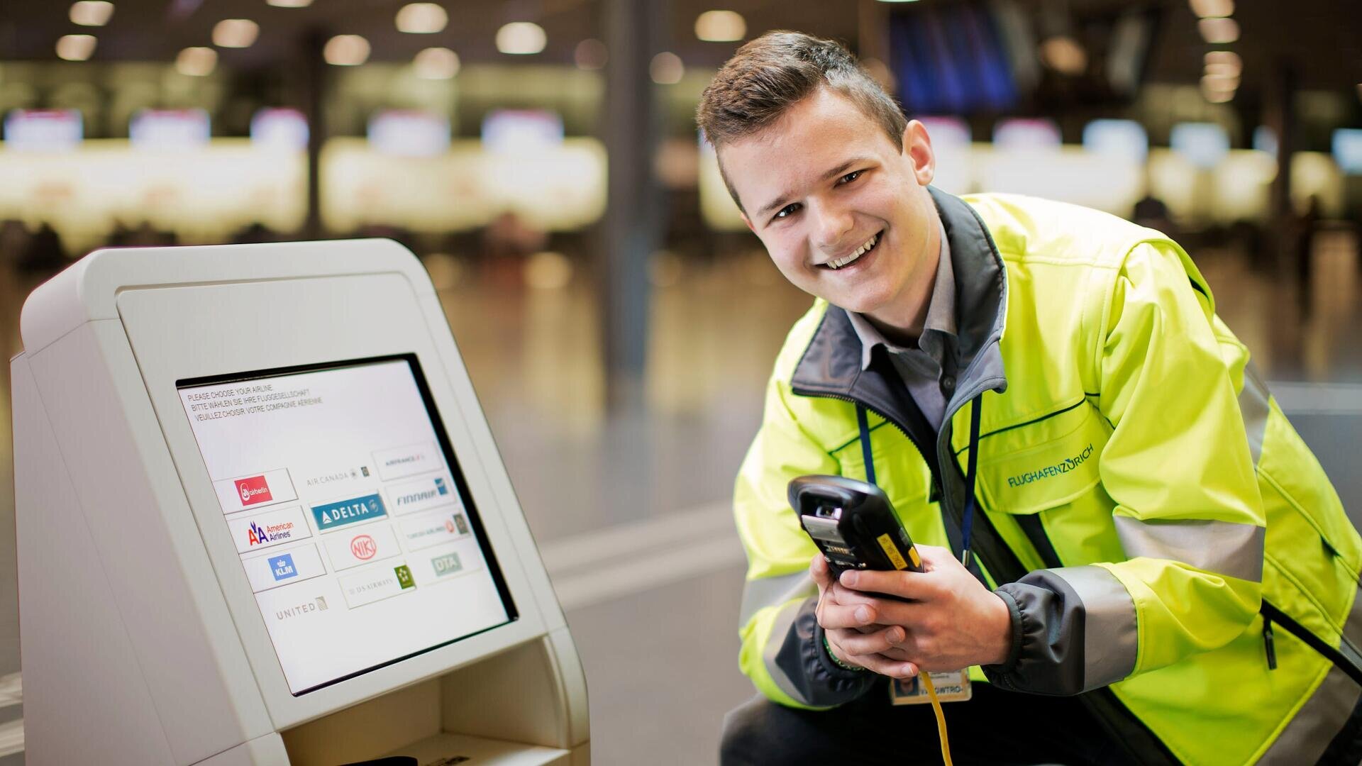 Ein lächelnder Flughafenmitarbeiter in einer gelben Jacke kniet neben einem Selbstbedienungs-Check-in-Kiosk und hält einen Scanner. Der Bildschirm des Kiosks zeigt die Logos verschiedener Fluggesellschaften. Im Hintergrund ist ein unscharfes Flughafenterminal zu sehen.