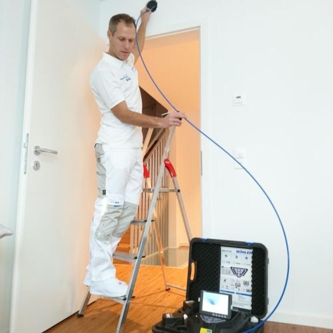 Eine Person in weißer Arbeitskleidung steht auf einer Stufenleiter und inspiziert mit einem blauen Kabel und einer Kamera die Innenseite einer Wand, während in der Nähe Geräte und ein kleiner Monitor auf dem Boden stehen.