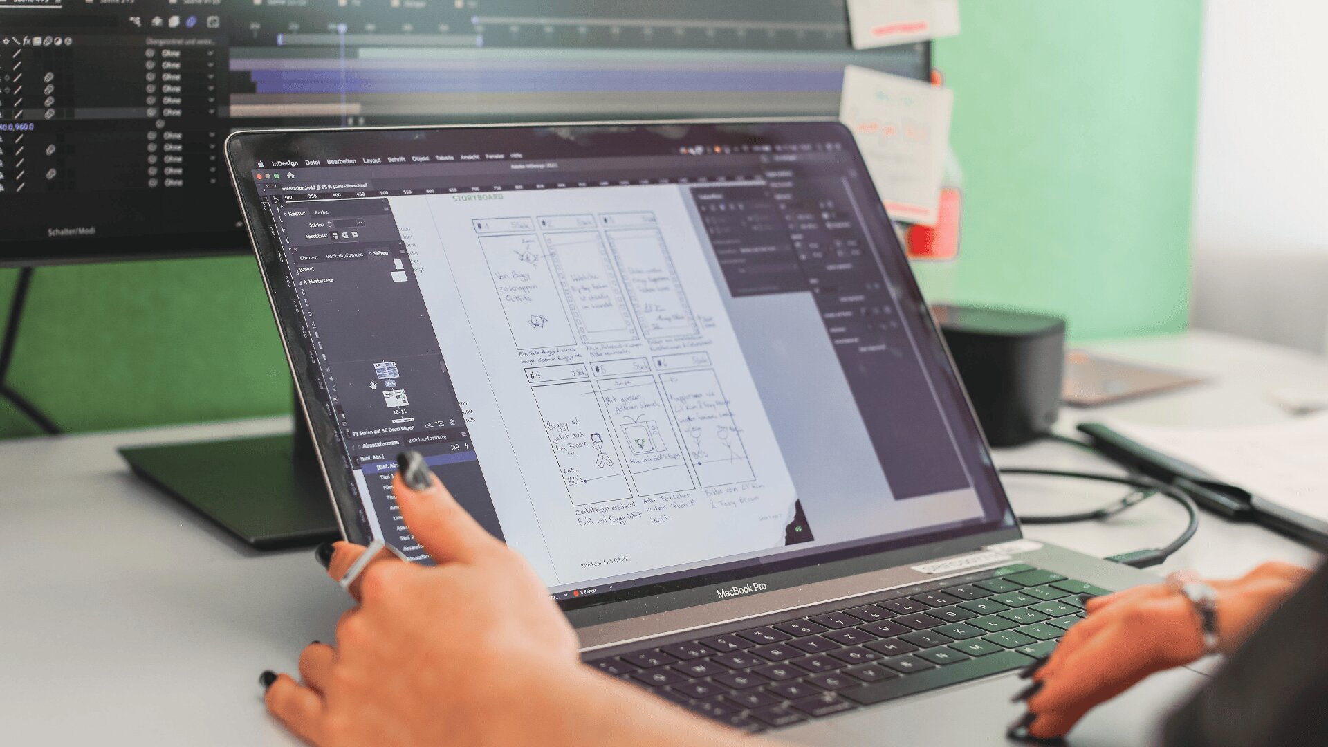 Eine Person arbeitet an einem Laptop, auf dem ein Wireframe-Design für eine mobile App angezeigt wird. Auf einem Monitor im Hintergrund ist eine Animationssoftware geöffnet, und der Arbeitsbereich ist mit Notizen und technischem Zubehör aufgeräumt.