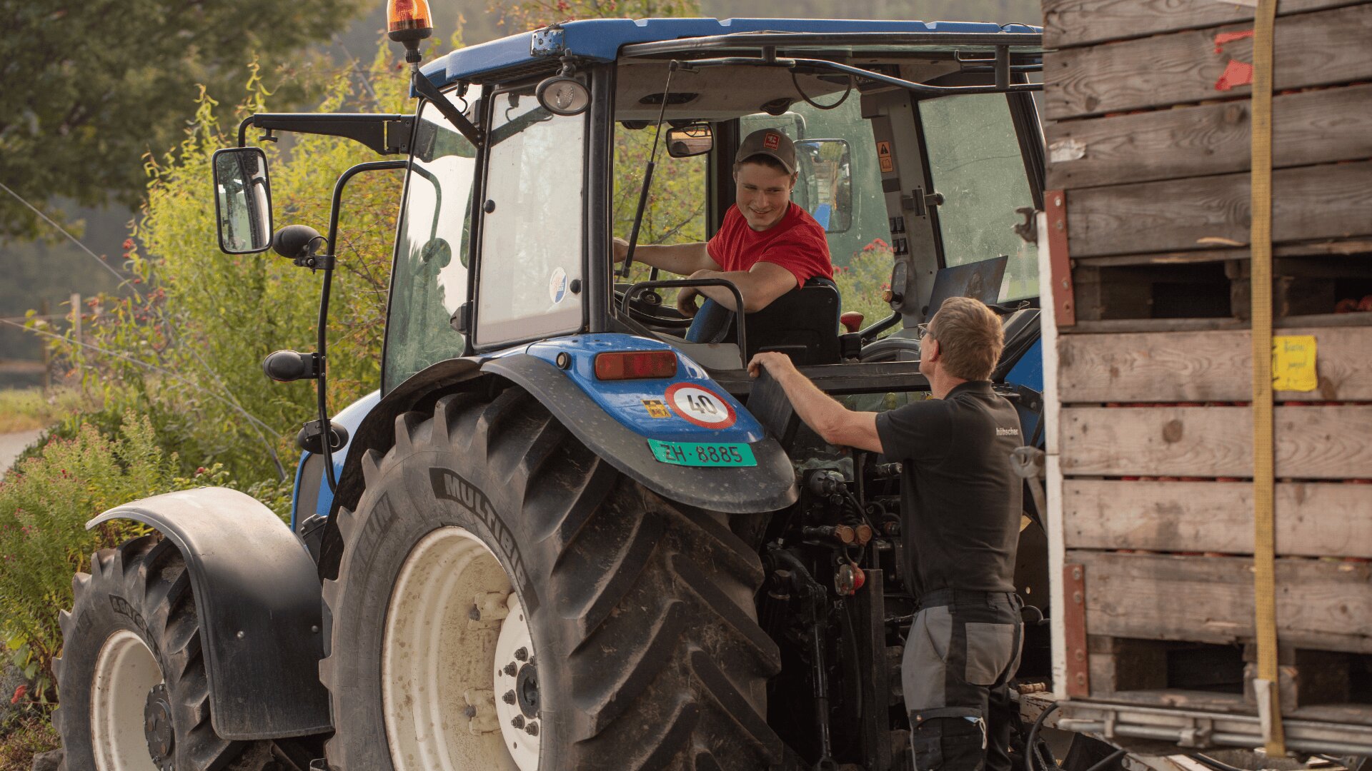 Zwei Personen unterhalten sich, während die eine in einem blauen Traktor sitzt und die andere daneben steht, in der Nähe einer Holzkiste. Die Szene spielt sich im Freien ab, mit Grünflächen im Hintergrund.