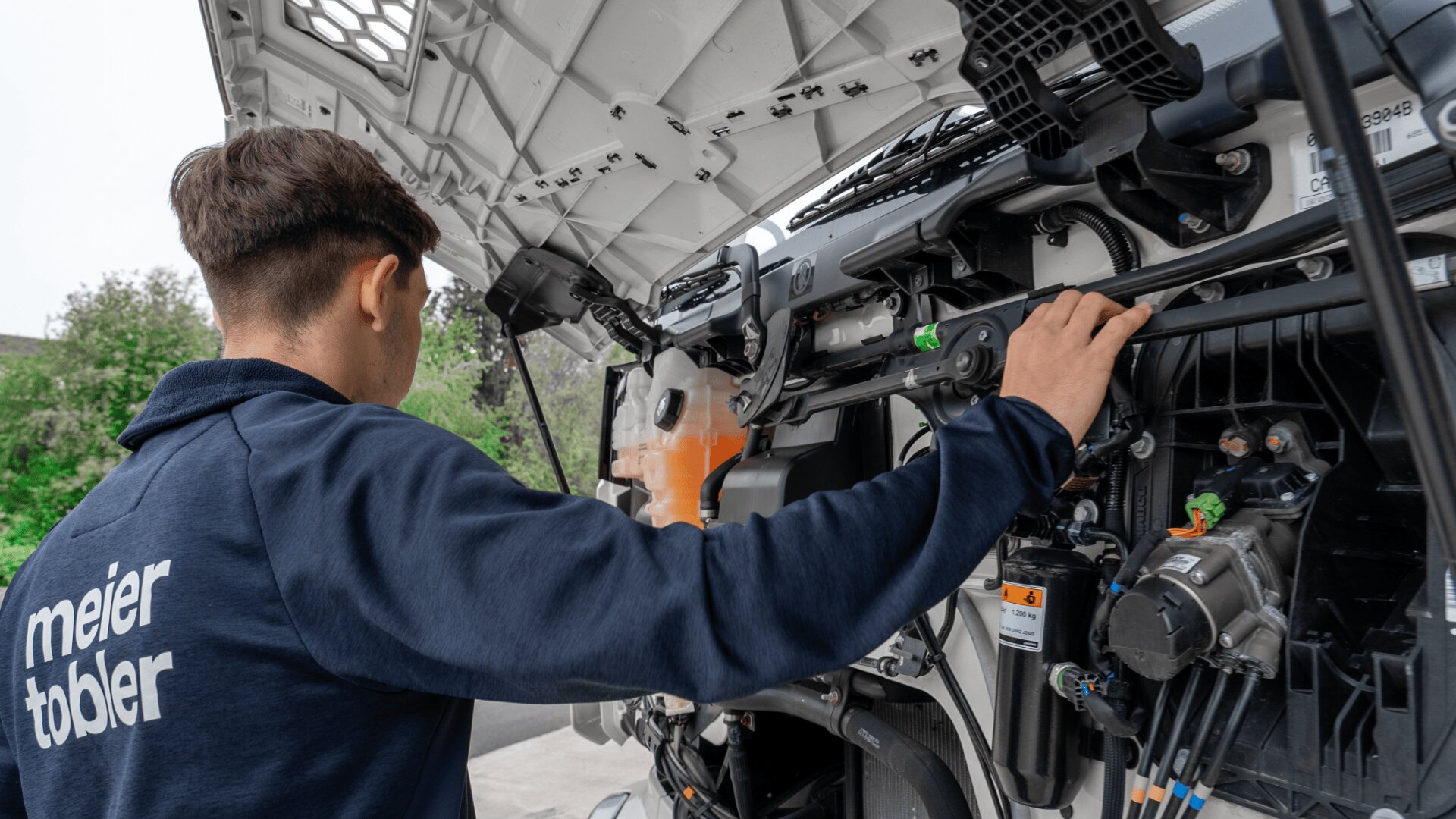 Ein Mechaniker, der eine Meier-Tobler-Jacke trägt, inspiziert den Motor eines großen Fahrzeugs bei geöffneter Motorhaube und stellt Komponenten ein, während er im Freien steht.