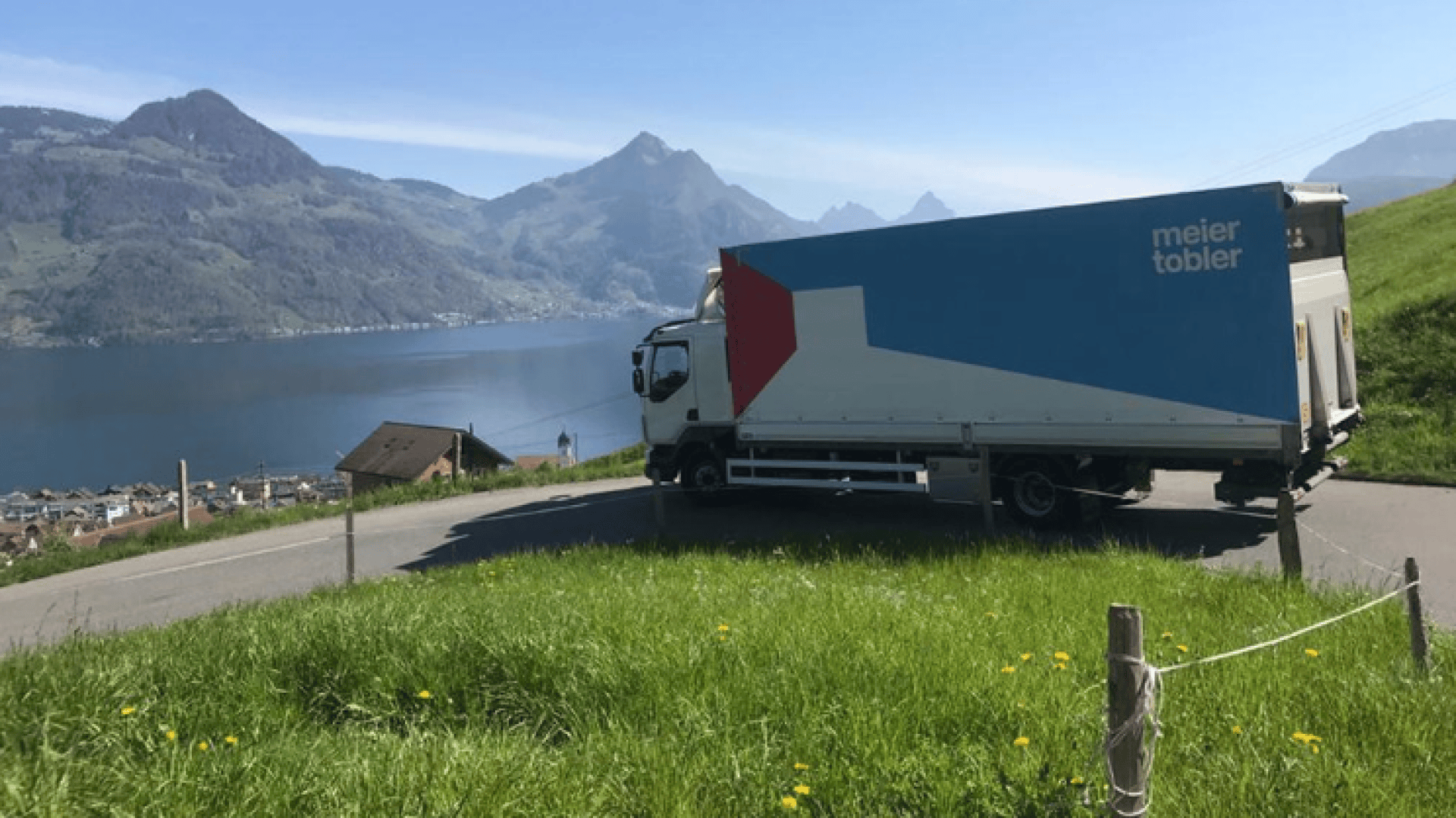Ein Lieferwagen von Meier Tobler fährt auf einer kurvenreichen Straße an einem See entlang, umgeben von grünen Hügeln und Bergen unter einem klaren blauen Himmel.
