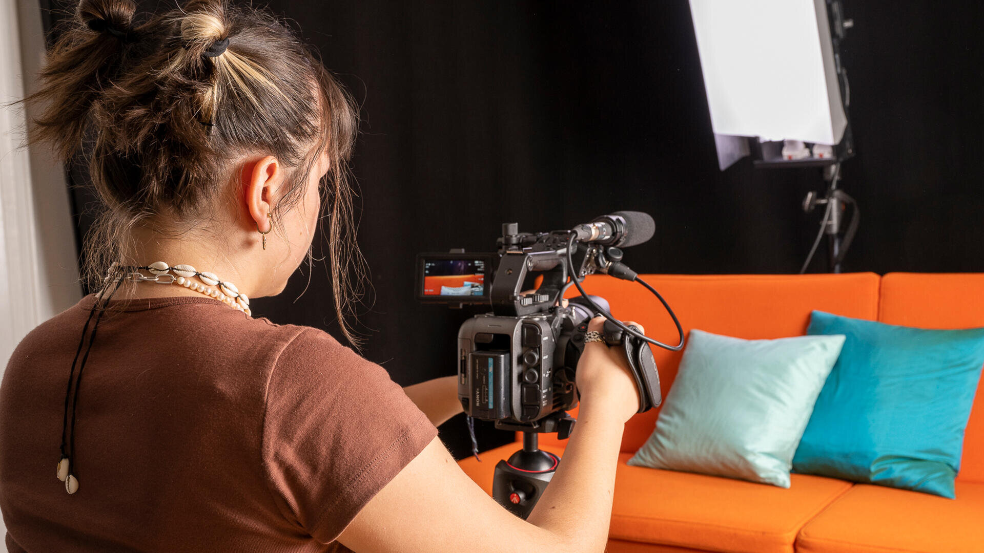 Eine Person mit braunen Haaren, die zu einem Dutt frisiert sind, filmt eine orangefarbene Couch mit blaugrünen und weißen Kissen mit einer professionellen Videokamera in einem Studio mit Studiobeleuchtung.