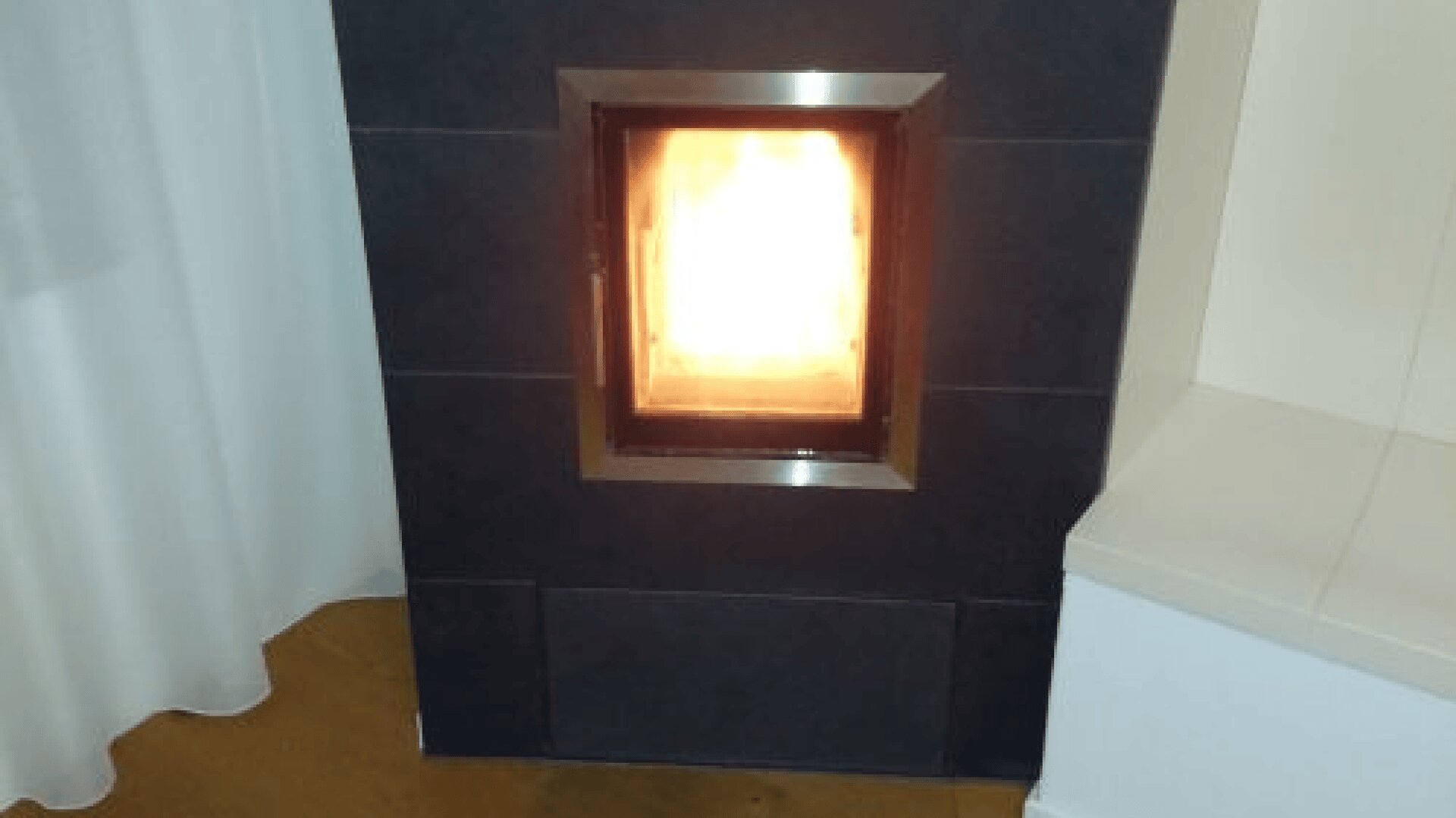 Ein beleuchteter Kamin mit einer hellen Flamme steht in einer dunklen, gekachelten Wand neben einem weißen Vorhang und einer weißen Sitzecke.