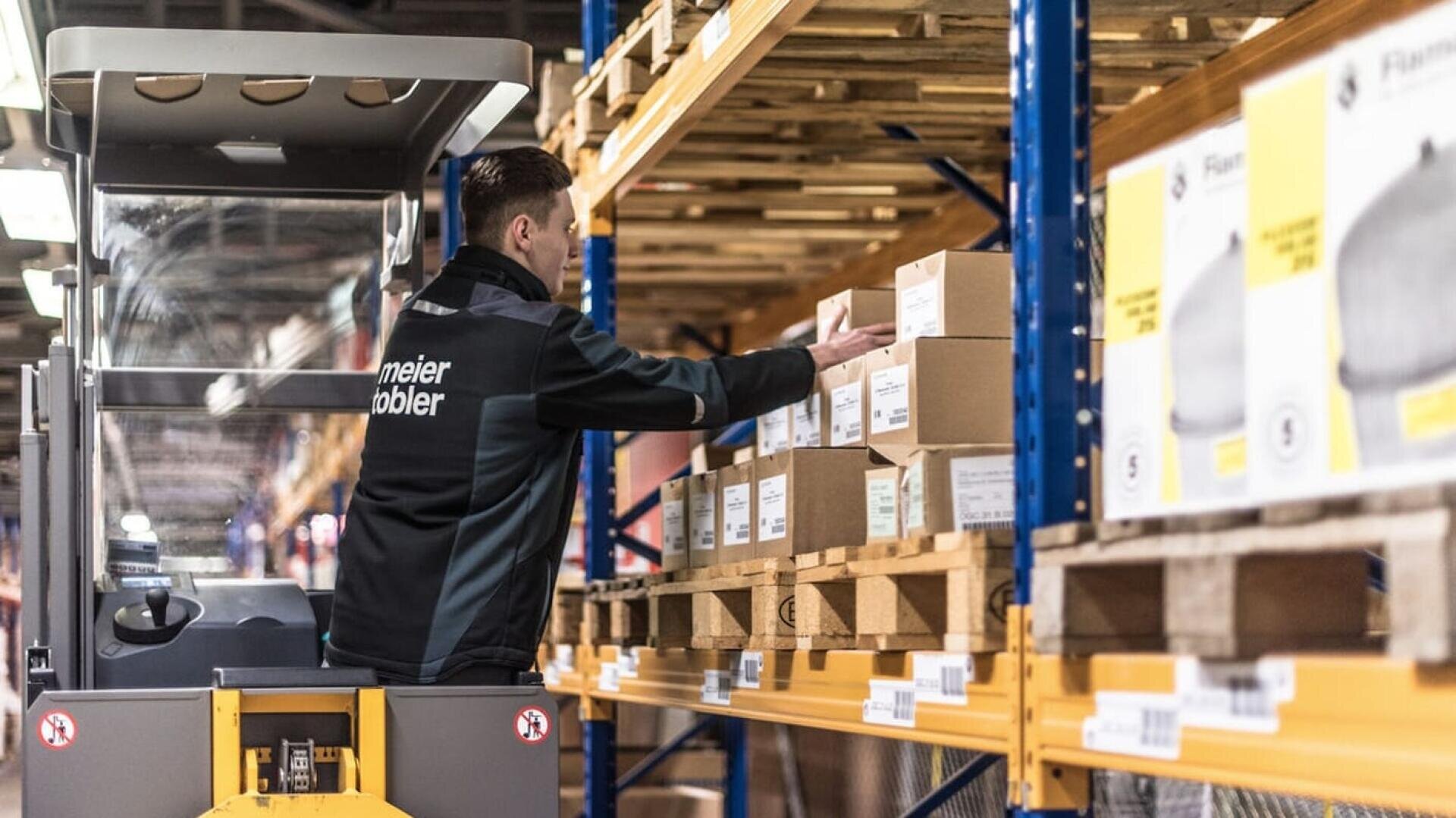 Ein Arbeiter bedient einen Gabelstapler und greift nach einem Karton in einem Lagerregal, umgeben von ordentlich gestapelten Kartons und gelben Produktetiketten.
