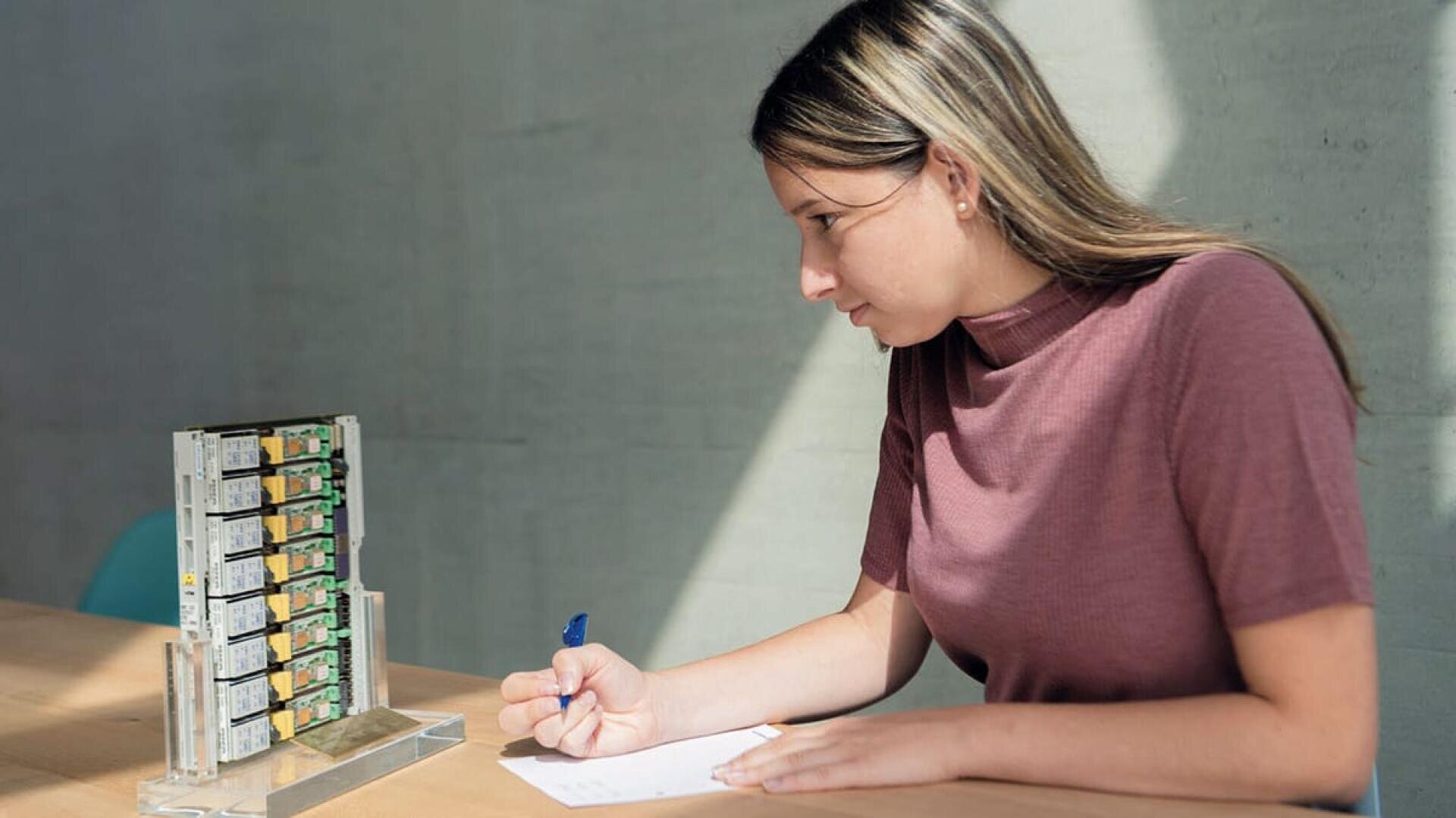 Eine Frau sitzt an einem Tisch und macht sich mit einem Stift Notizen auf Papier, während sie aufmerksam auf ein kleines, vertikales Regal mit Computer-Hardware-Komponenten auf einem Ständer schaut.