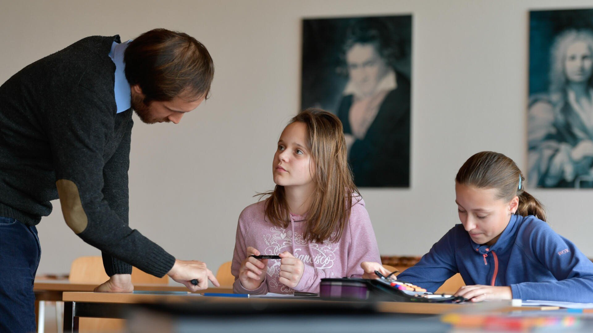 Eine Lehrerin beugt sich vor, um zwei Mädchen zu helfen, die an einem Schreibtisch in einem Klassenzimmer sitzen. Ein Mädchen hört aufmerksam zu, während das andere schreibt. An der Wand im Hintergrund hängen Porträts von historischen Persönlichkeiten.