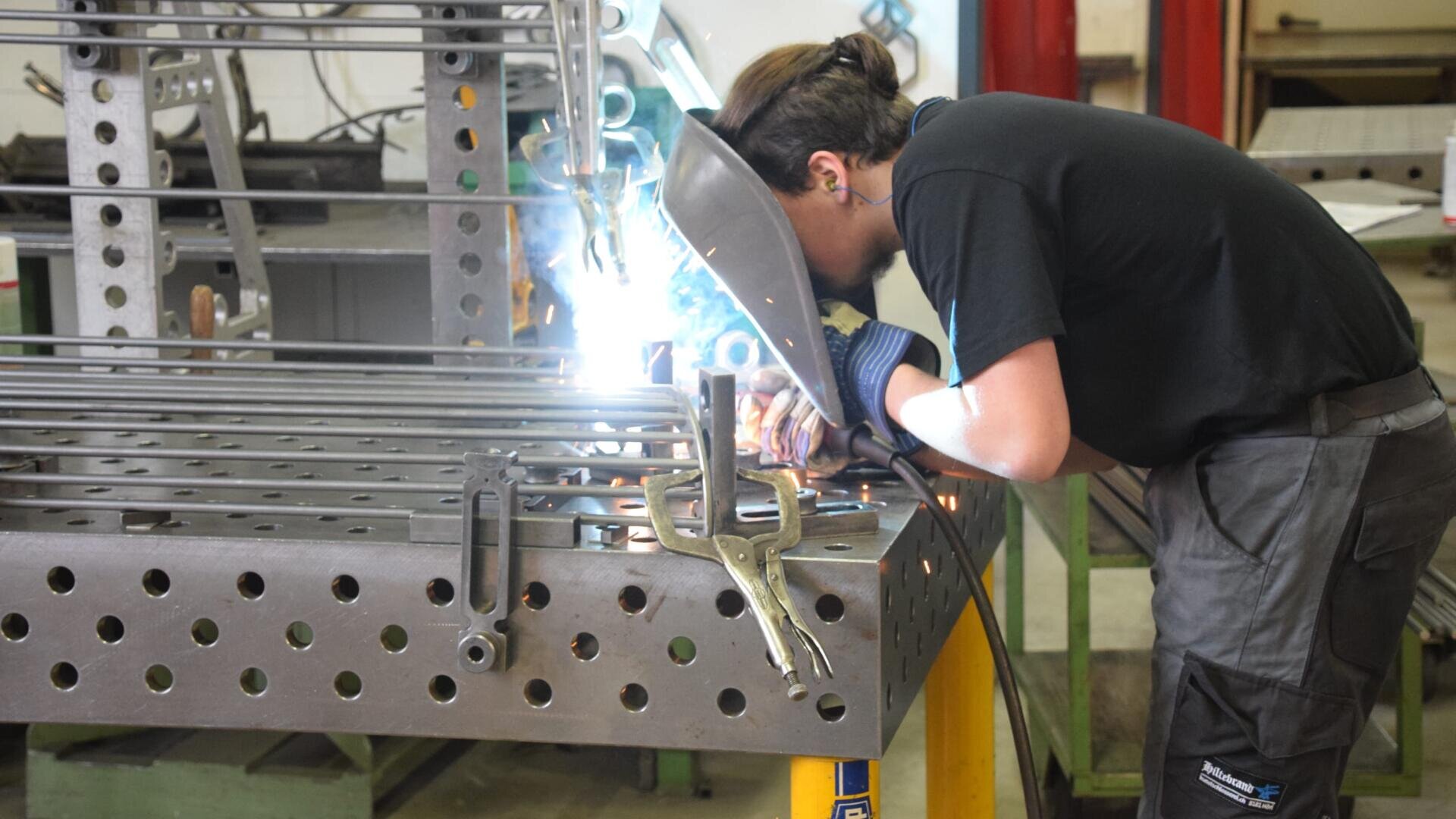 Eine Person in Schutzkleidung schweißt in einer Werkstatt Metallstangen auf einem perforierten Arbeitstisch, wobei Funken und helles Licht vom Schweißvorgang sichtbar sind.