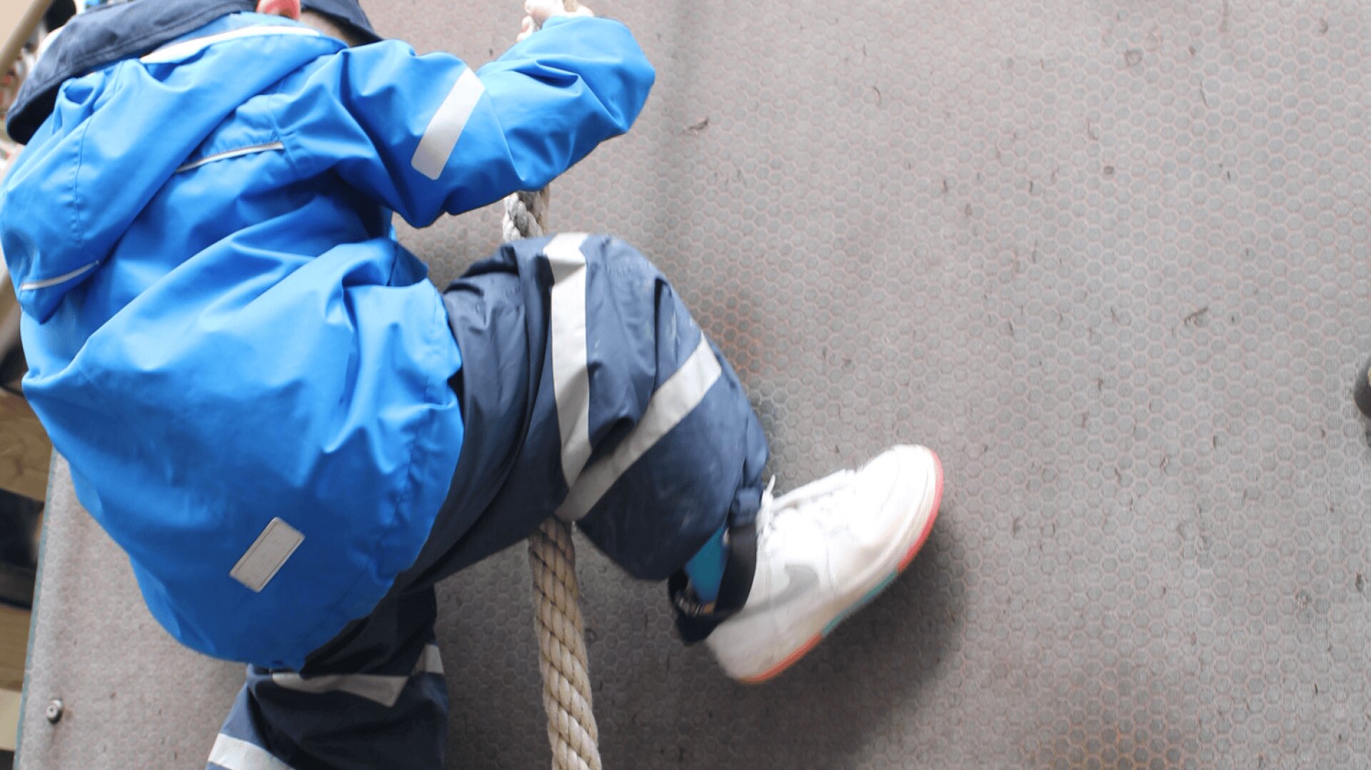 Ein Kind in einer blauen Jacke und einer reflektierenden Hose klettert an einem Seil über eine graue Fläche, wobei ein weißer Turnschuh zu sehen ist.
