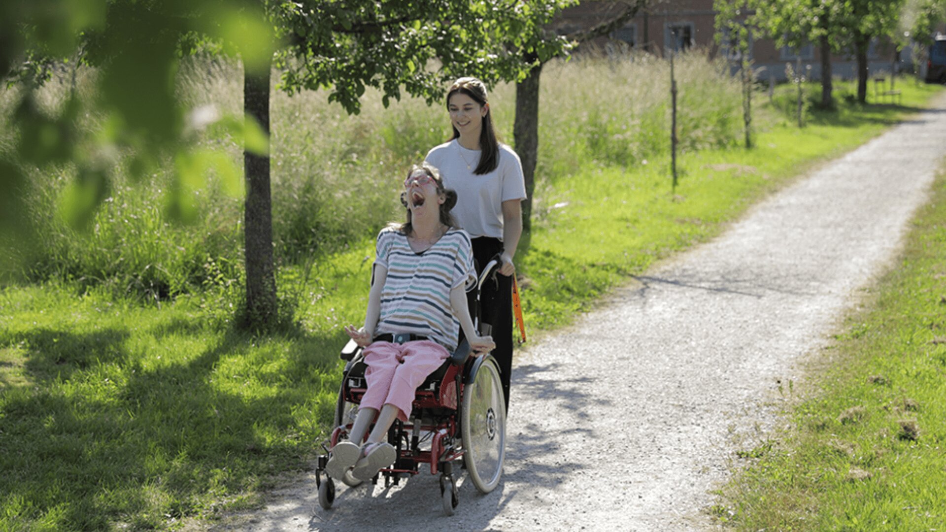 Eine junge Frau schiebt eine andere Frau im Rollstuhl über einen sonnigen, von Bäumen gesäumten Weg. Beide lächeln und genießen die freie Natur. Der Weg ist von grünem Gras und hohen Bäumen umgeben.