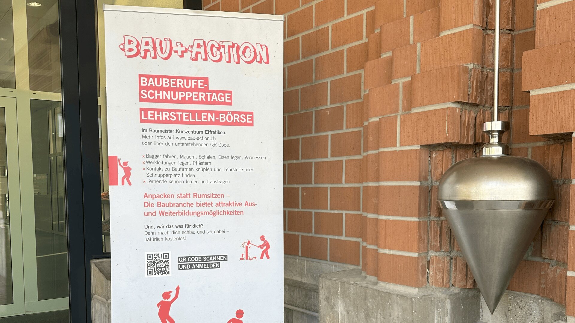 Ein stehendes Banner in deutscher Sprache, das für die Ausbildung bei Bau+Action und die Karrieretage im Baugewerbe wirbt, ist in der Nähe einer Backsteinmauer neben einem großen Metalllot angebracht. Das Banner ist mit rotem Text und Symbolen zum Thema Bauwesen versehen.