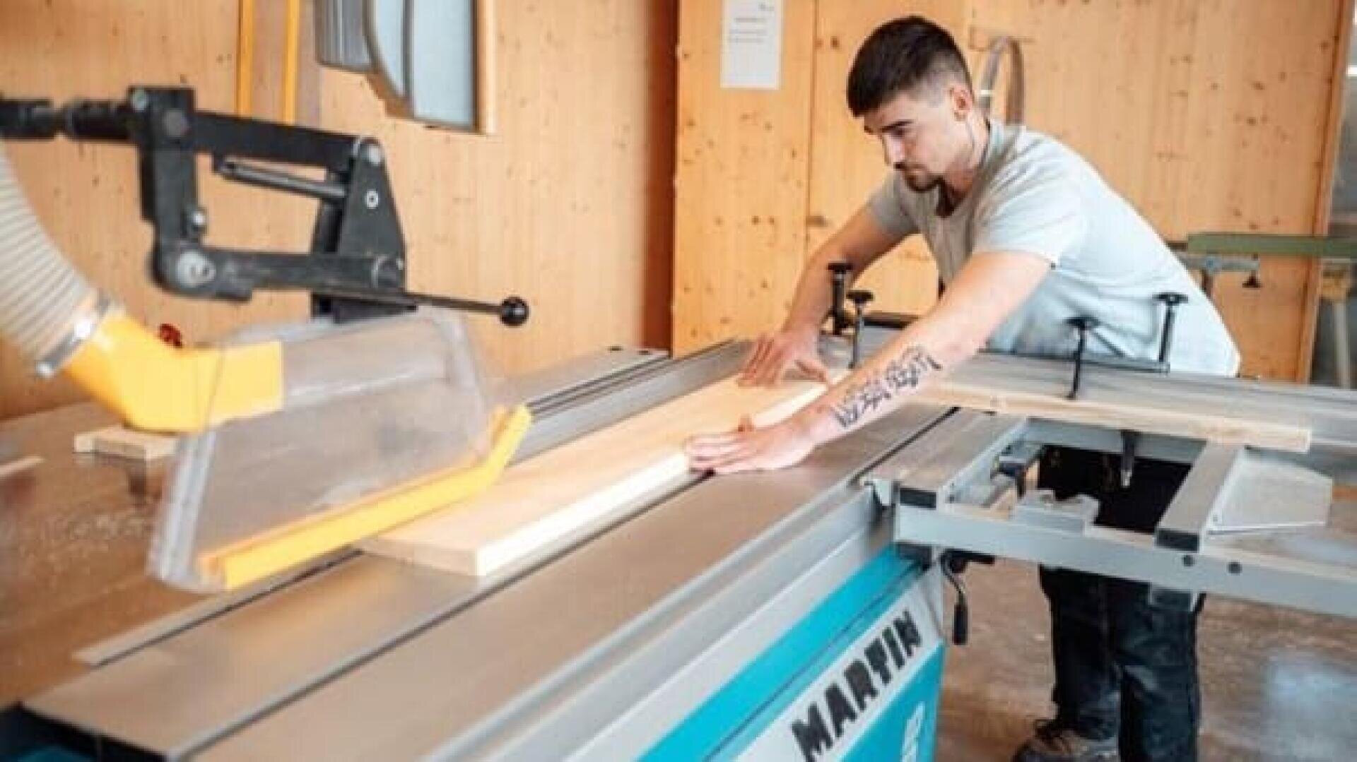 Ein Mann schneidet mit einer Tischsäge ein Holzbrett in einer Werkstatt. Er trägt ein hellgraues Hemd und führt das Holz mit beiden Händen, um Präzision zu gewährleisten.